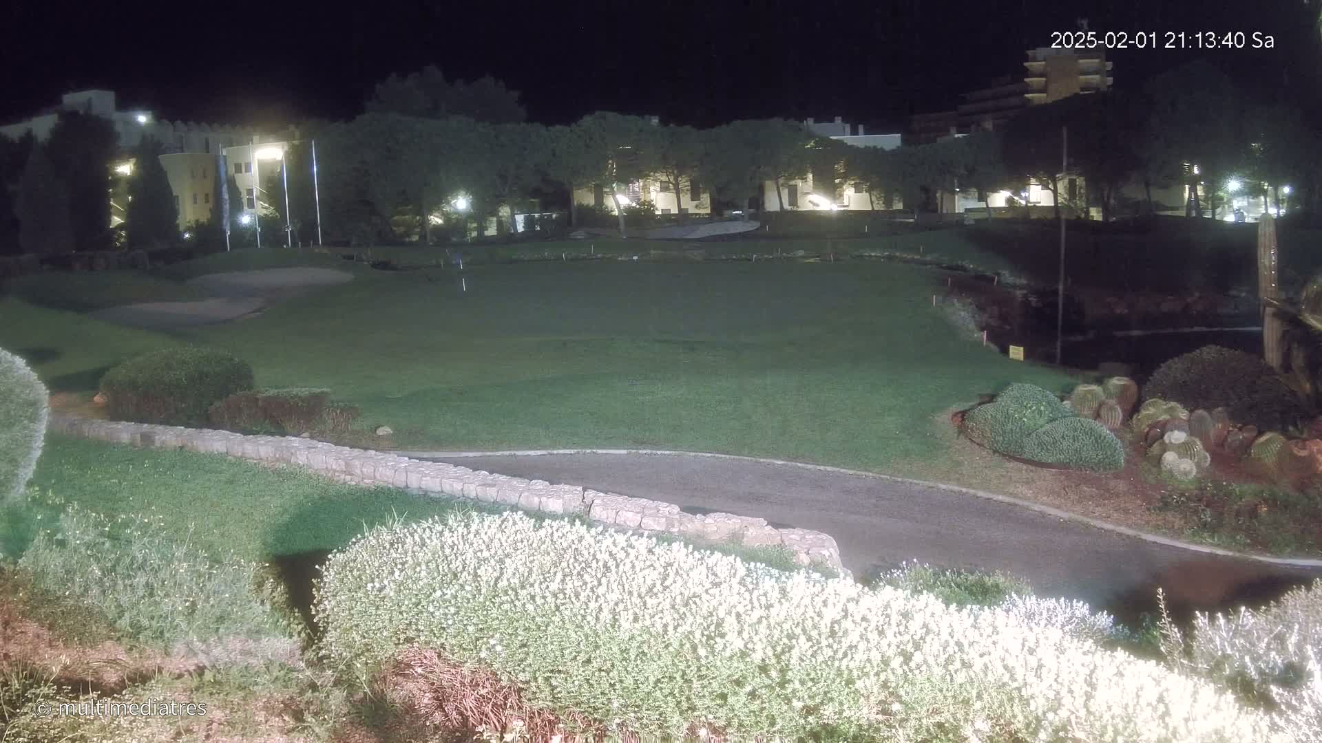 A nighttime view of a grassy putting green, bordered by low stone walls and flowering shrubs, with a building and trees in the background under artificial lighting.
