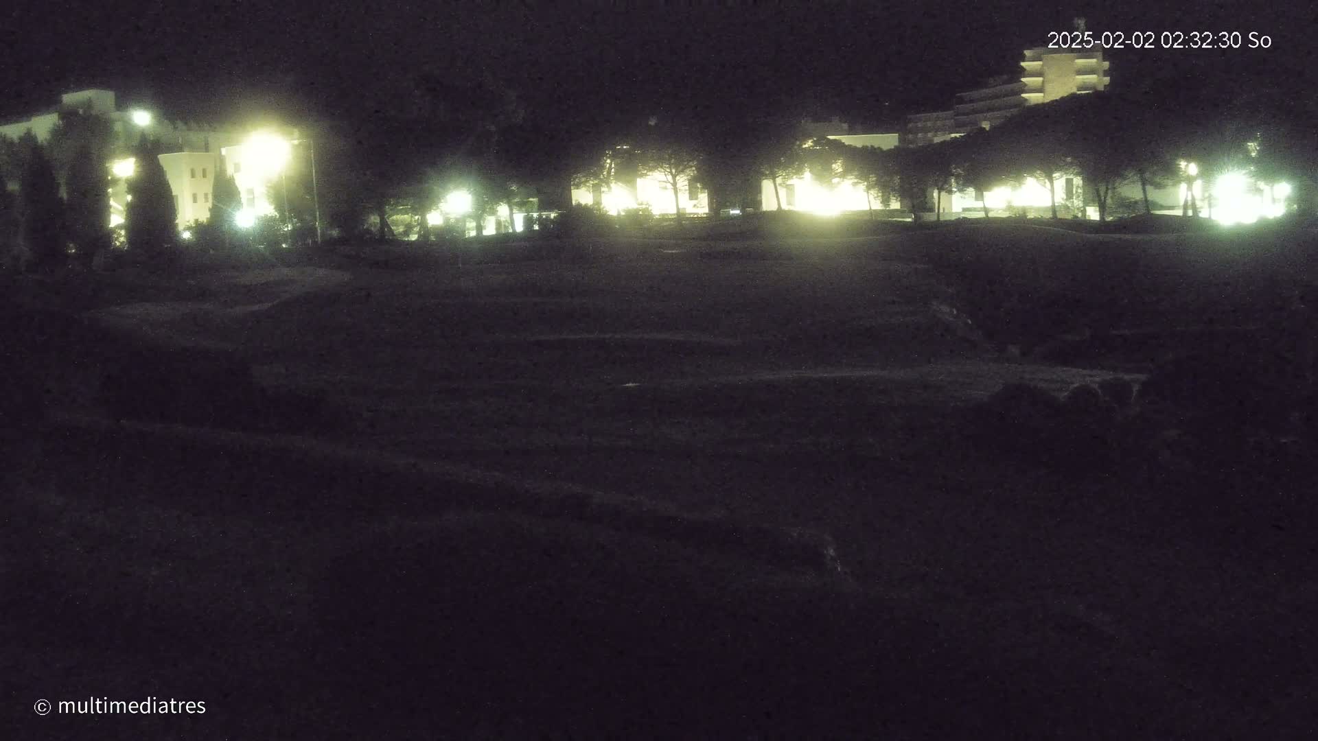 A dark, nighttime scene shows illuminated buildings behind a line of trees, overlooking a mostly flat, dark expanse of land.