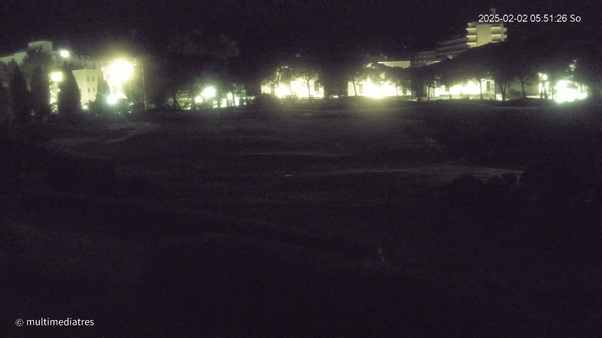 Illuminated buildings are visible at night behind a dark, grassy area.