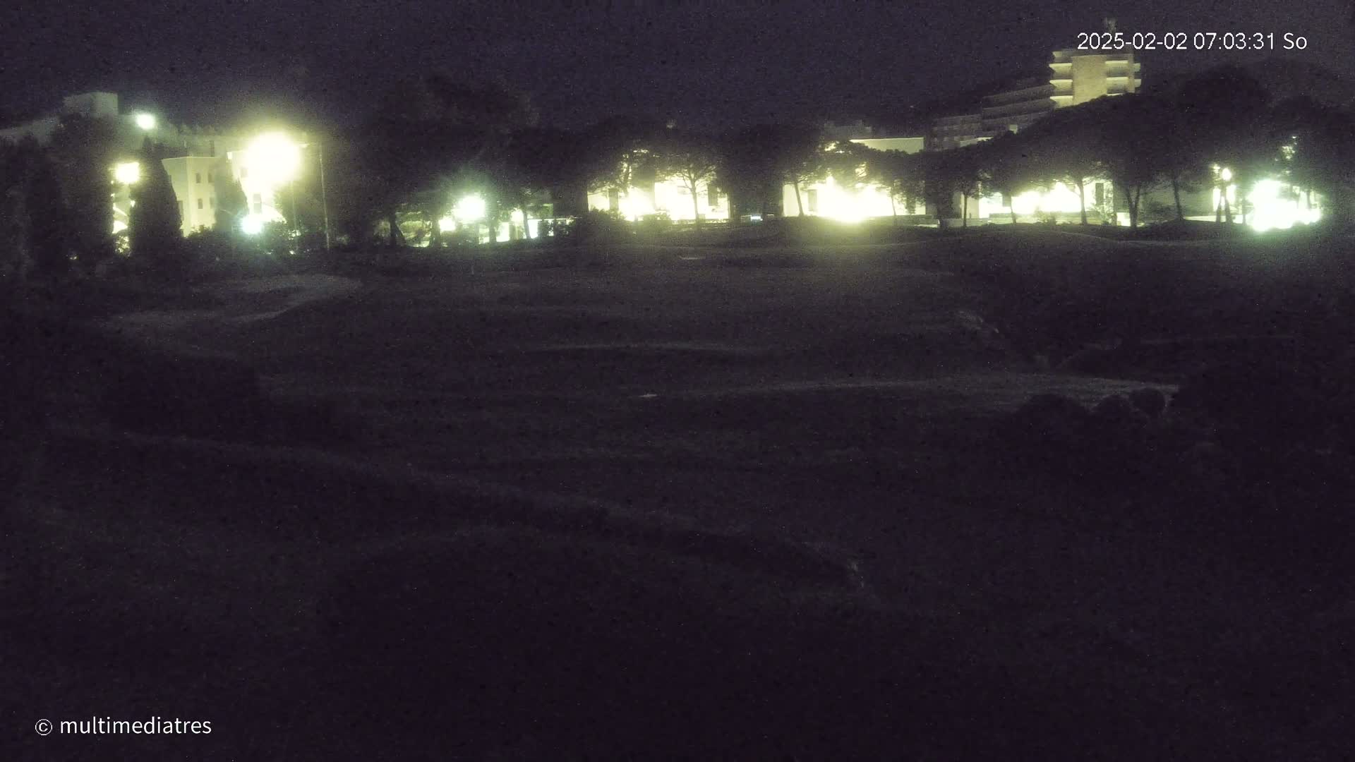 At night, illuminated buildings are visible behind a dark, grassy area.