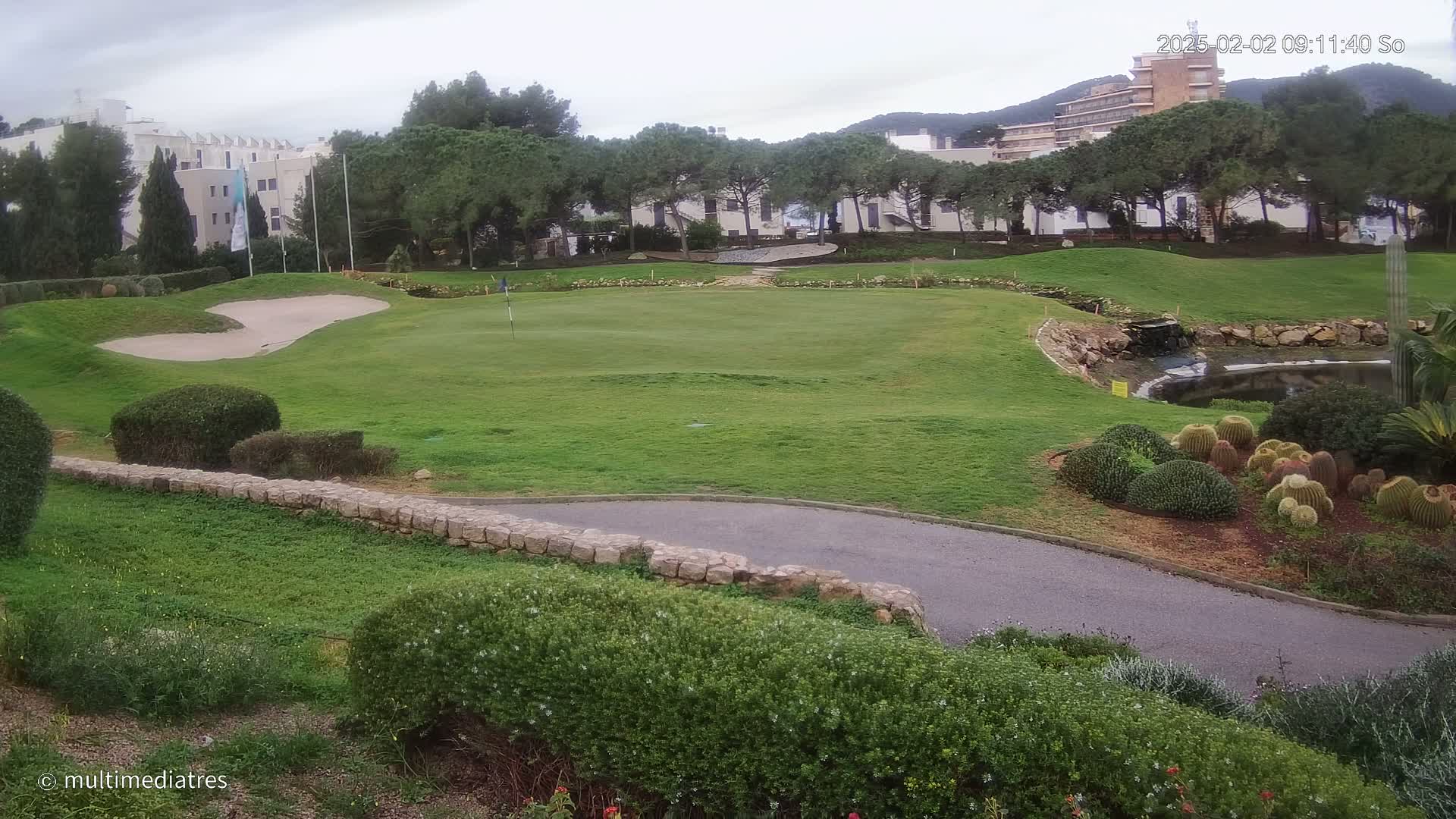 A golf course green with a sand trap and small pond is visible under an overcast sky, surrounded by shrubs, trees, and buildings in the background.
