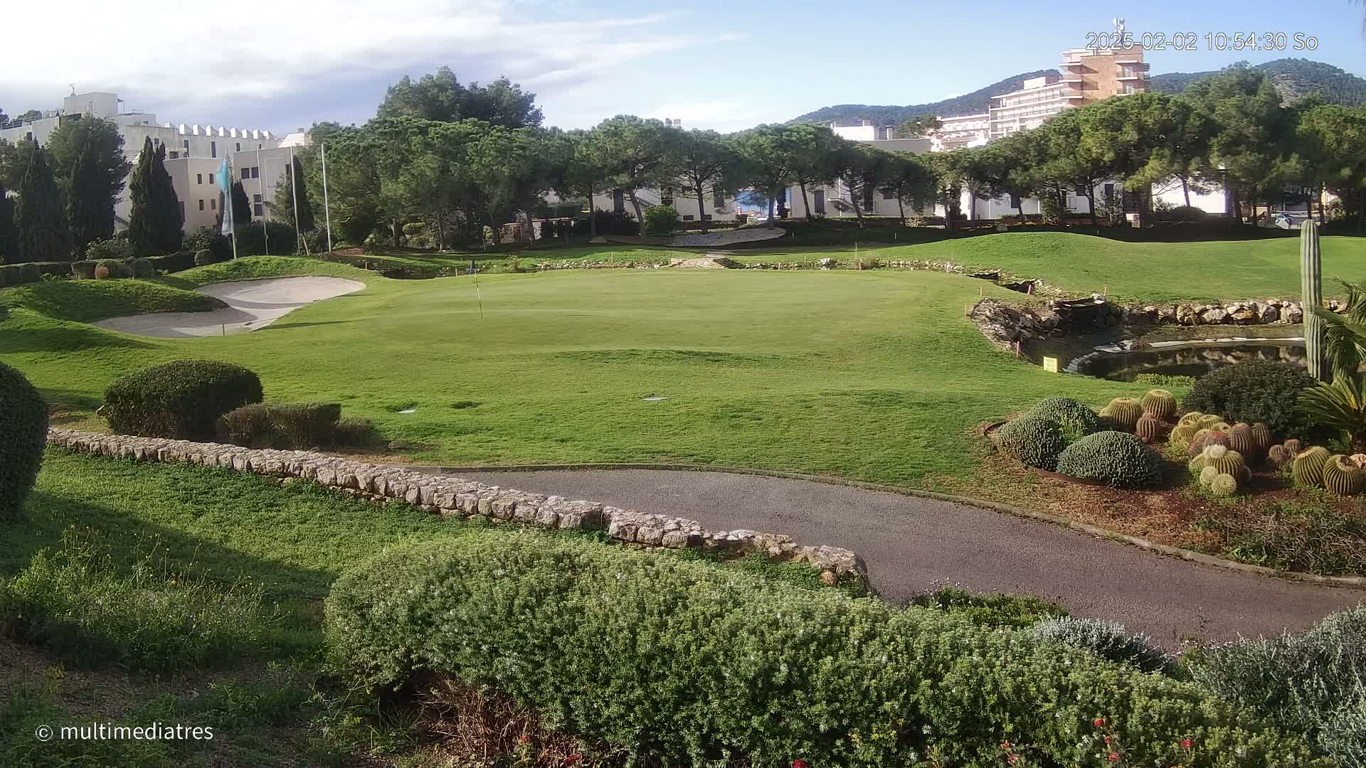 A golf course green with sand traps, a small pond, and cacti, situated near a building complex under a partly cloudy sky.