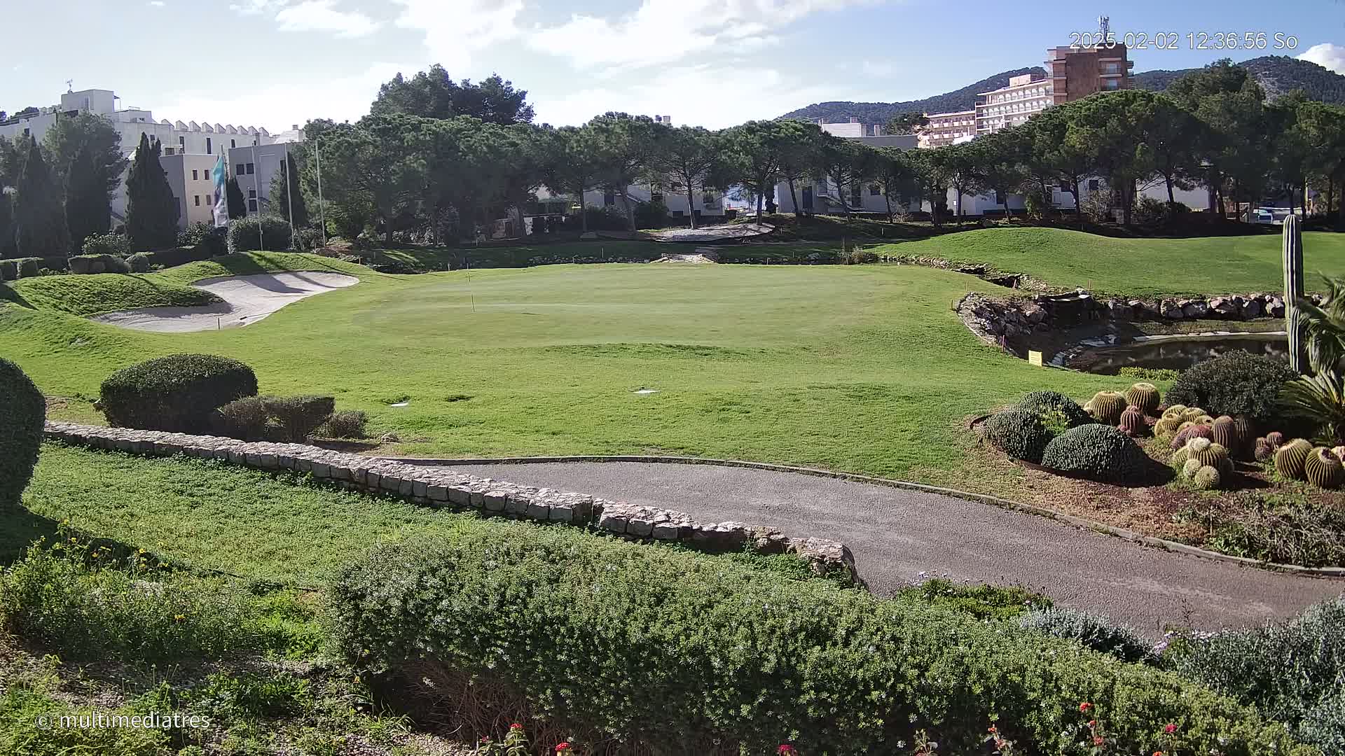 A sunny day on a golf course shows a green with a sand trap, a paved path, landscaping including cacti, and buildings in the background.
