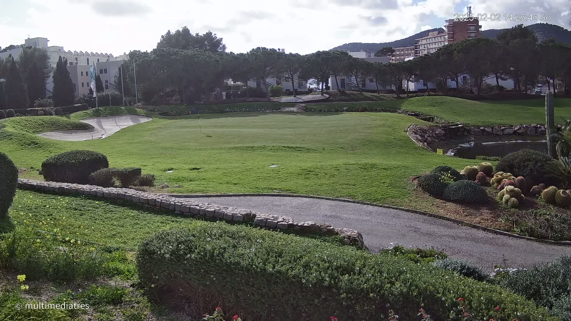 A putting green on a golf course, surrounded by landscaping and buildings, under partly cloudy skies.