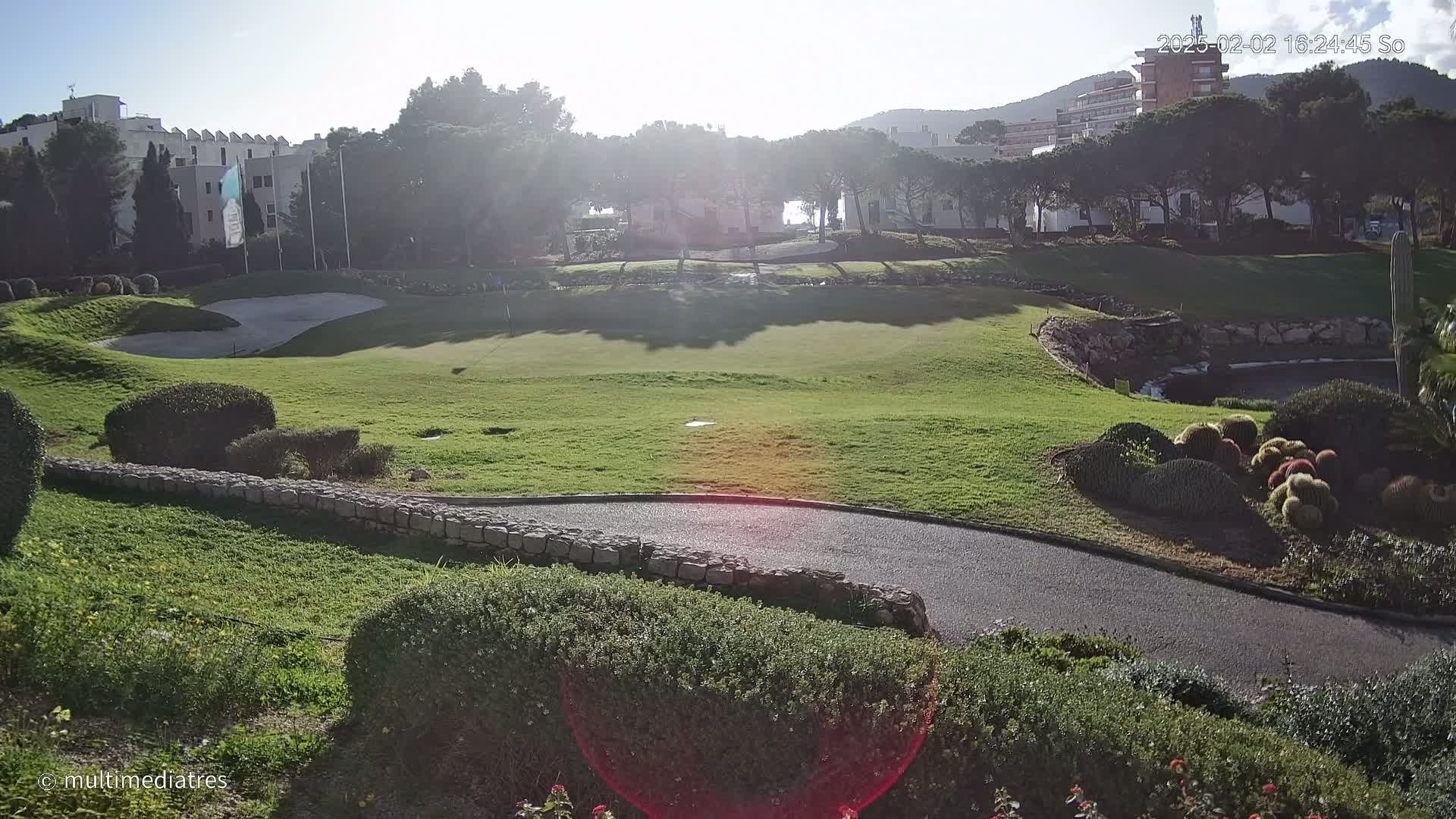 A sunny golf course with a green, sand trap, and a paved path winds through the manicured landscaping.