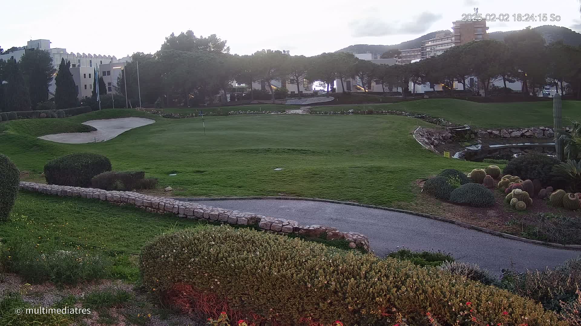 A golf course green with a sand trap, bordered by shrubs and a low stone wall, overlooks a paved path, a small pond, and cacti under a partly cloudy, dusky sky.