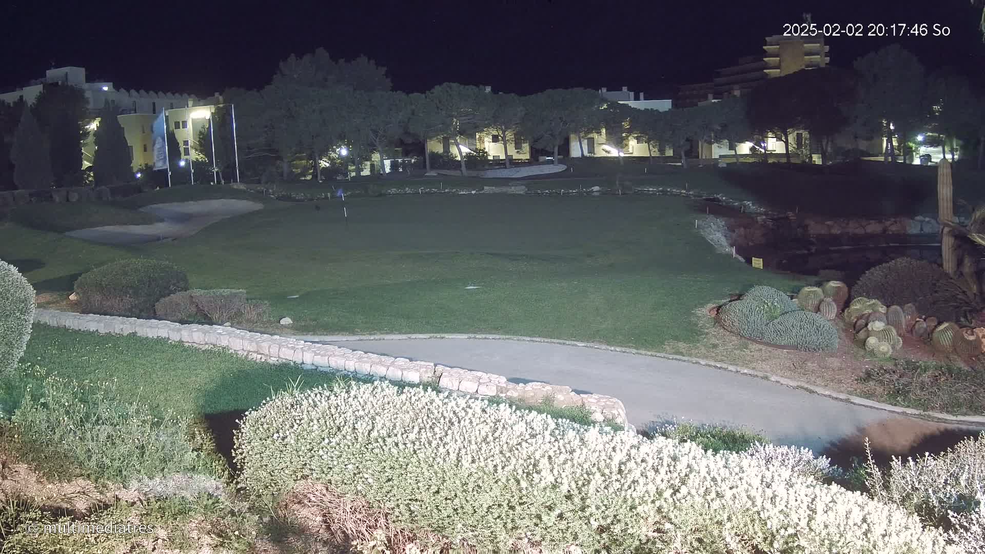 A nighttime view of a putting green, bordered by shrubs and a low stone wall, with a building and trees visible in the background under a dark sky.