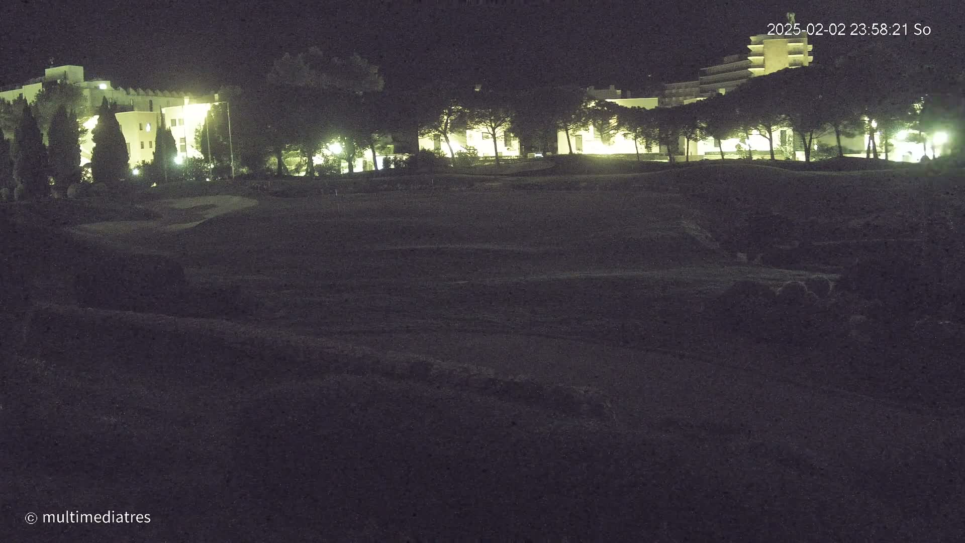 A nighttime view of a grassy area with illuminated buildings and trees in the background under a dark sky.