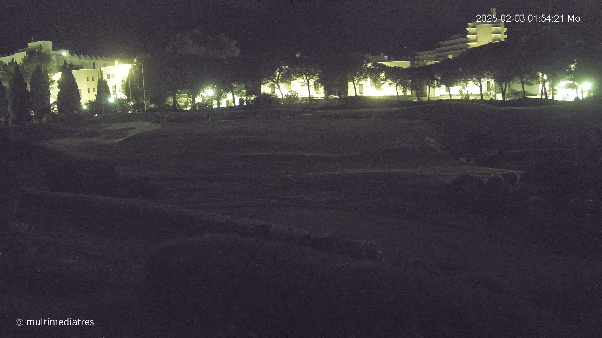 A nighttime view of a golf course with a few illuminated buildings in the background under a clear sky.