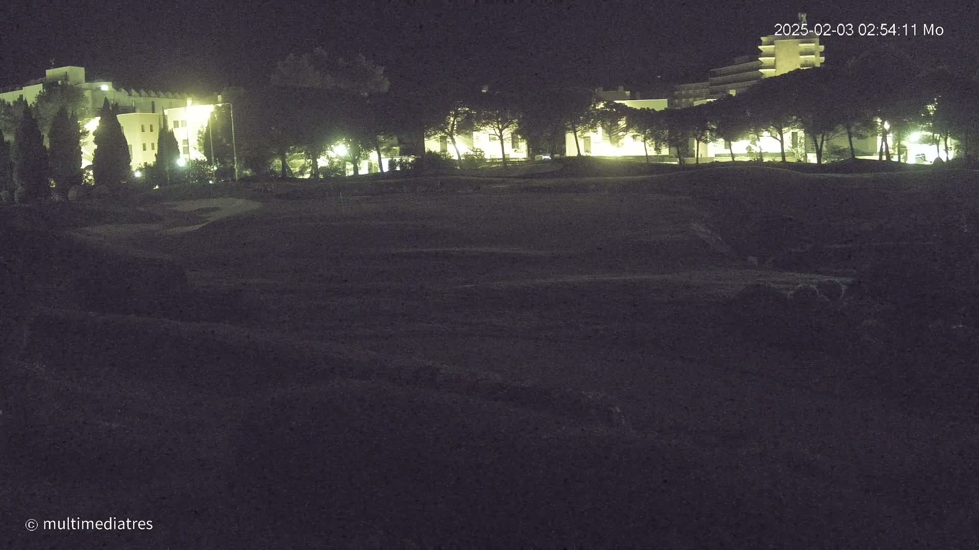 A nighttime view of a golf course with illuminated buildings and trees in the background under a dark sky.