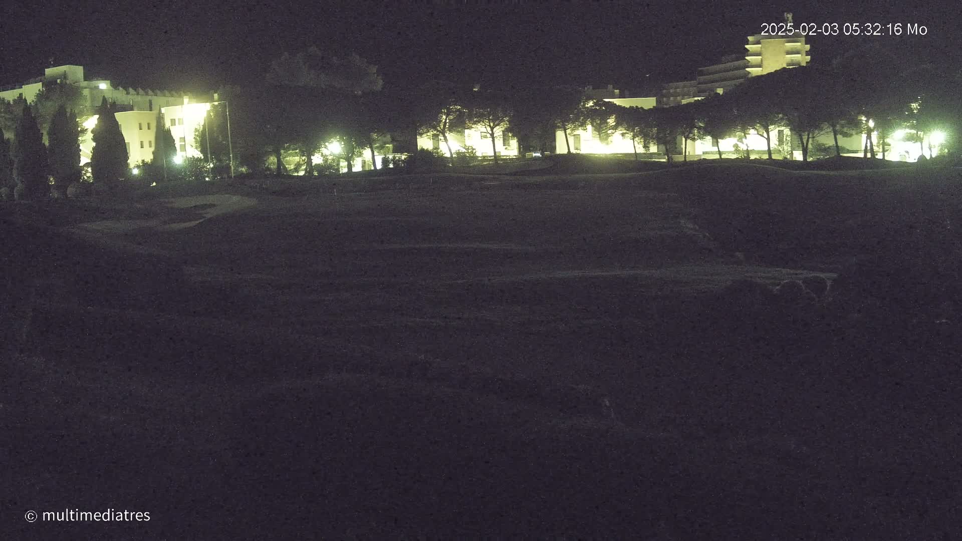 A dark, nighttime scene shows a grassy area in front of illuminated buildings surrounded by trees.