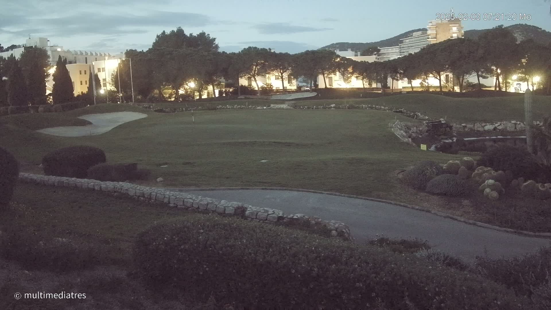A twilight view of a golf course with buildings in the background under a partly cloudy sky.