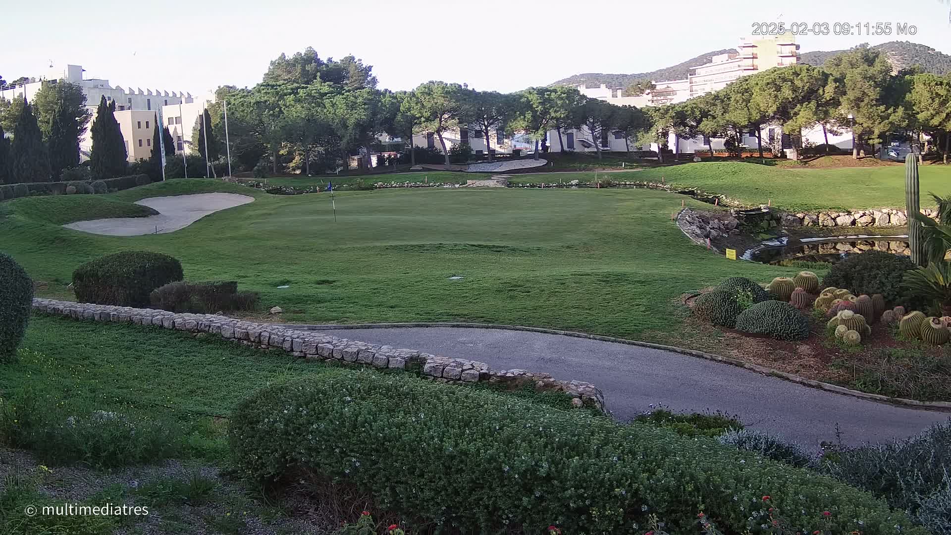 A golf course green with a sand trap, a small pond, and a low stone wall, surrounded by lush vegetation and buildings in the background under a clear sky.