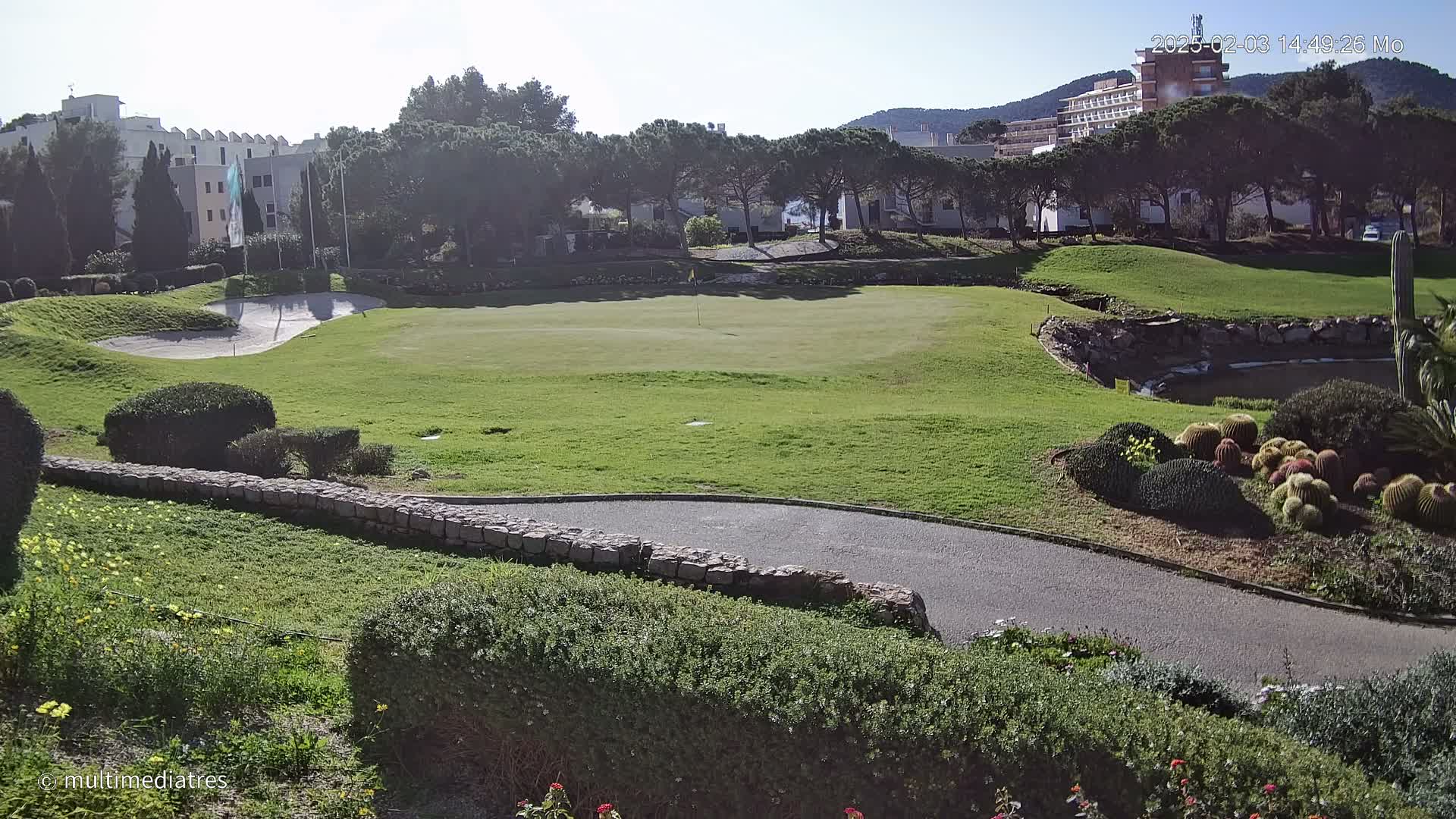 A sunny view of a putting green on a golf course, bordered by landscaping, a low stone wall, and buildings in the background.