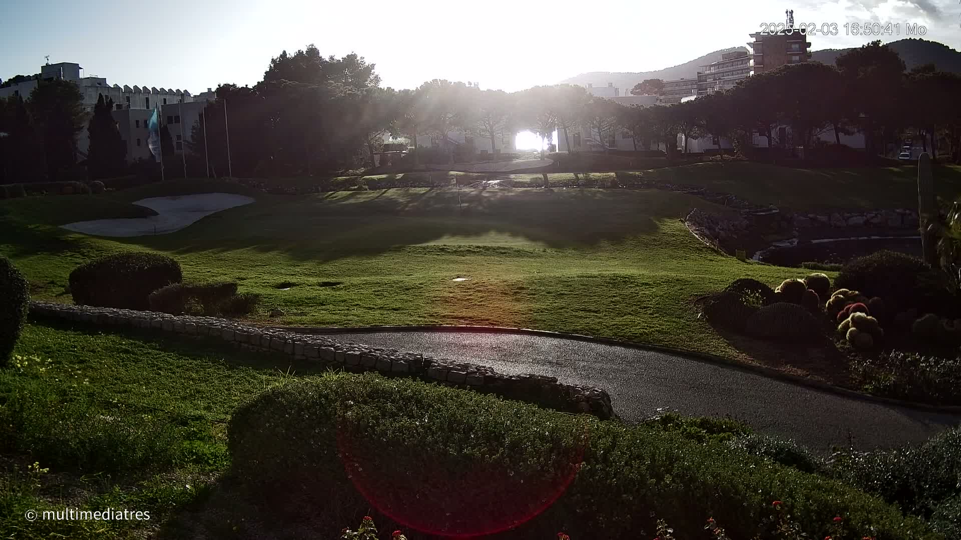 A sunlit golf course with a paved path winds through green grass, shrubs, and a stone wall, with buildings and trees in the background under a mostly clear sky.