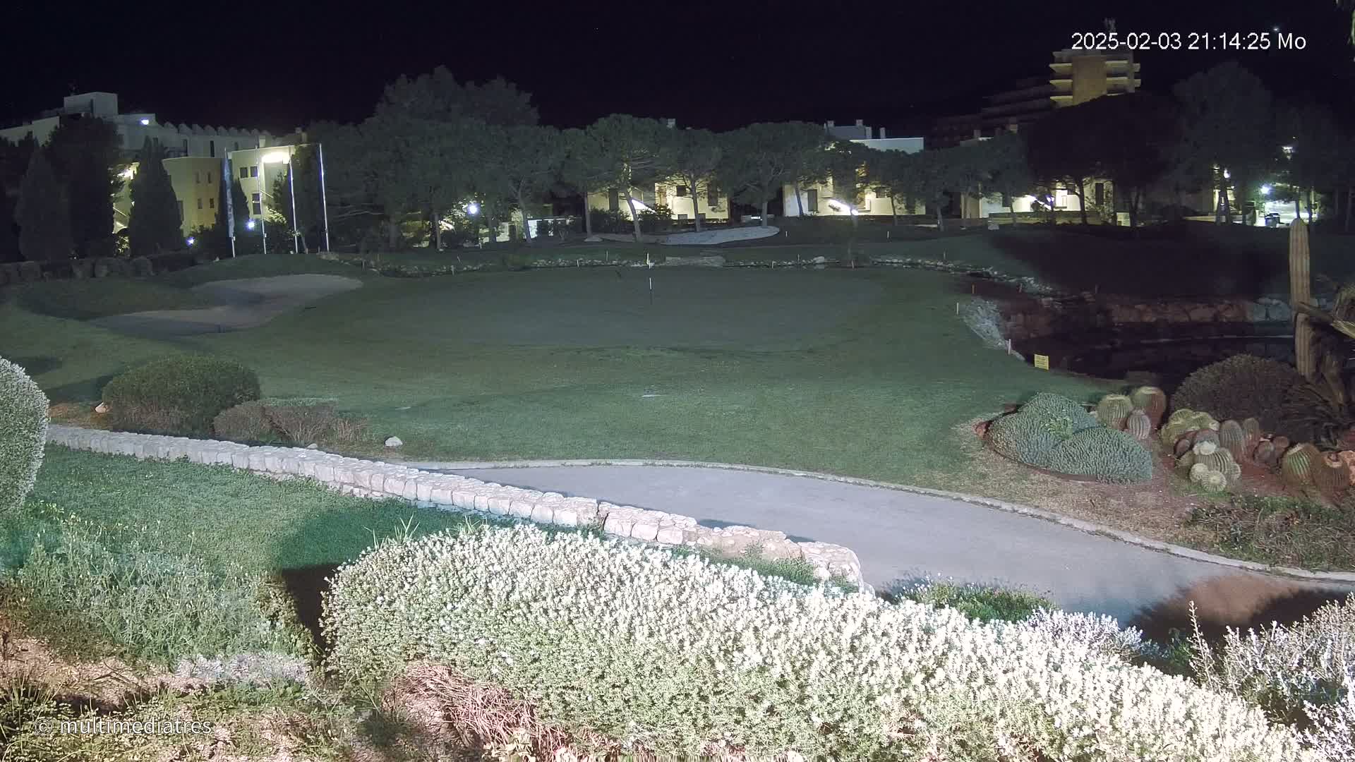 A nighttime view of a putting green next to a low stone wall, with buildings and trees illuminated in the background.