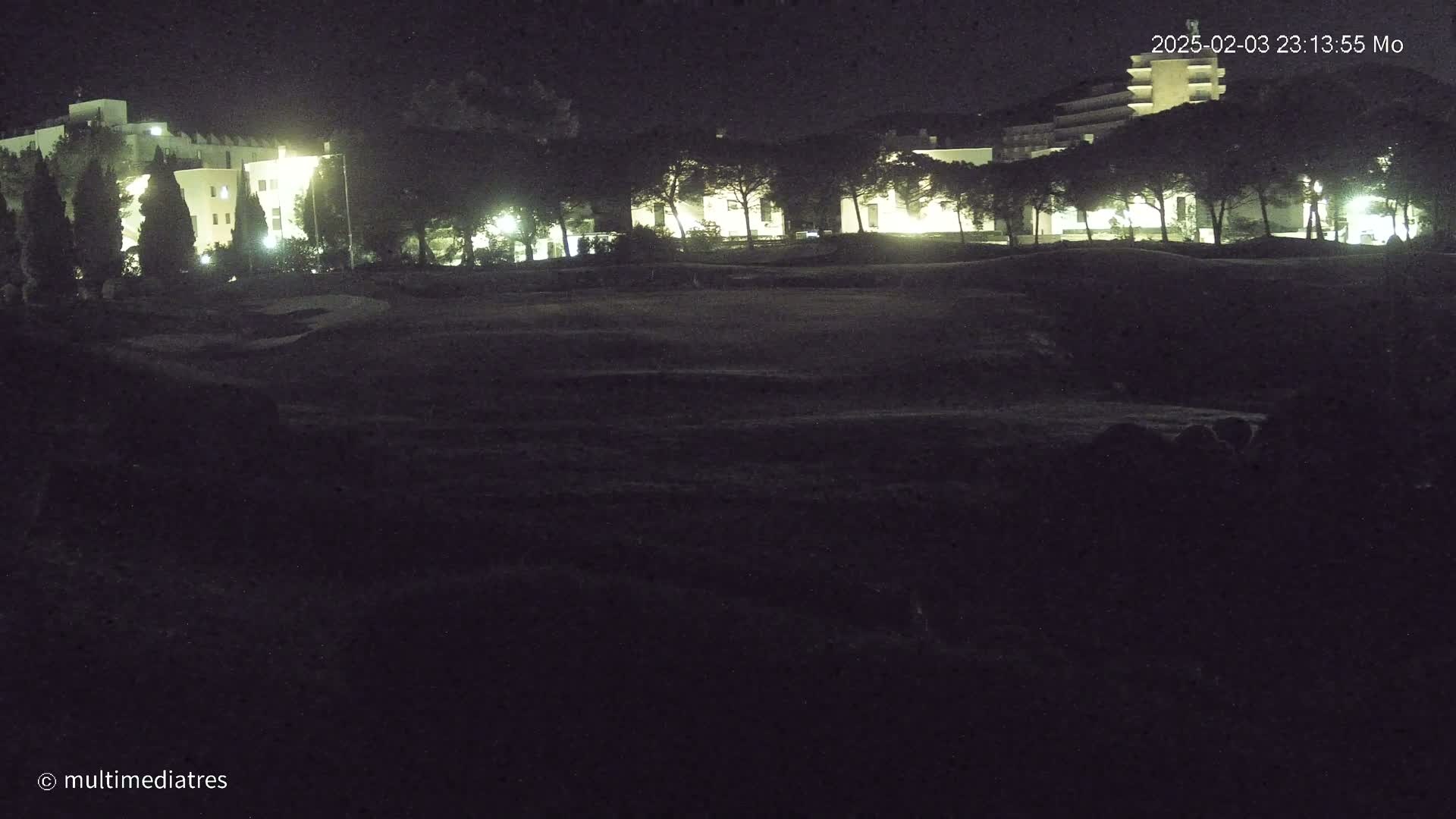 At night, illuminated buildings are visible behind a dark, grassy area.
