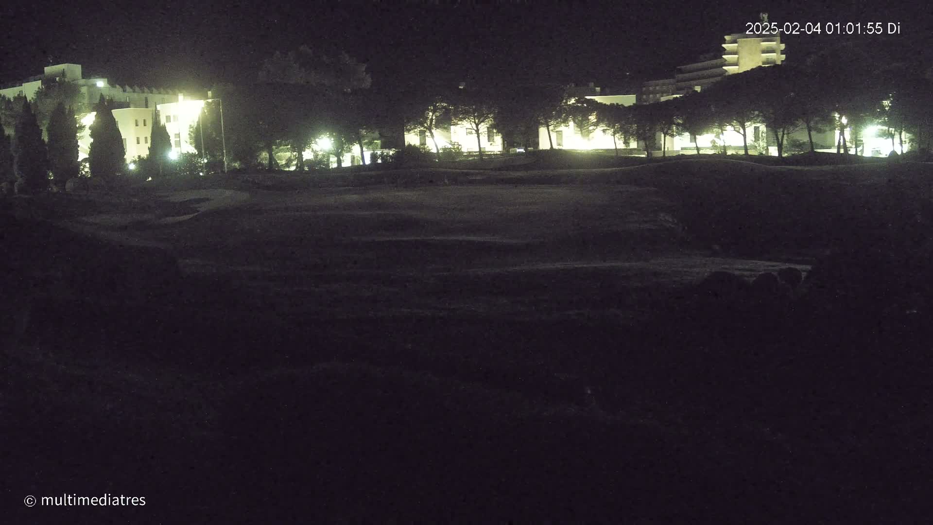 Lit-up buildings are visible behind a dark, grassy area at night.