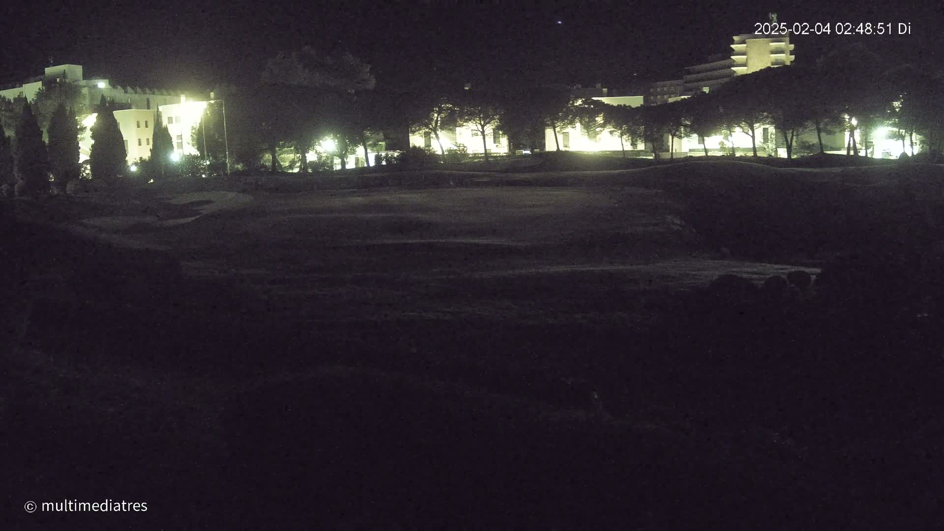 A nighttime view of a grassy area with illuminated buildings and trees in the background under a clear, dark sky.