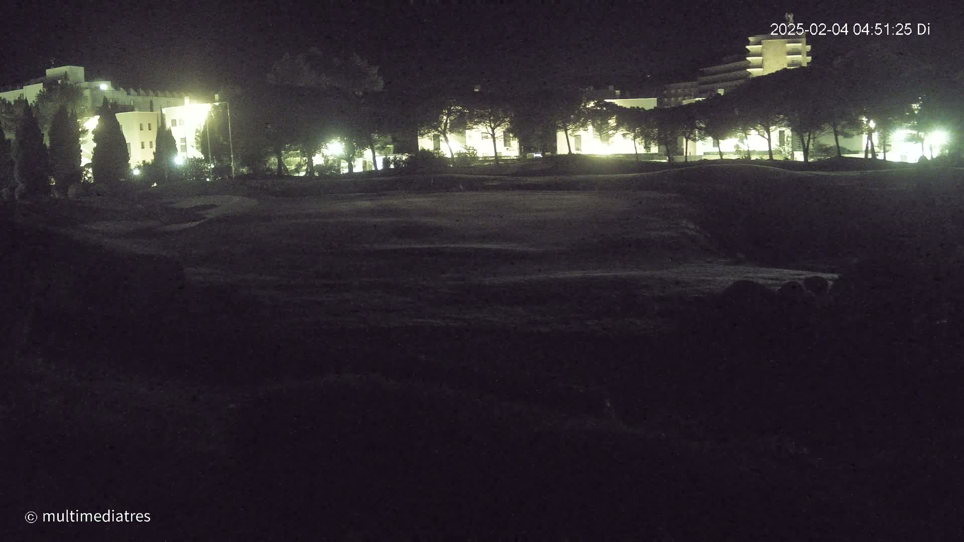 A nighttime view of dimly lit buildings and trees behind a dark, grassy area.