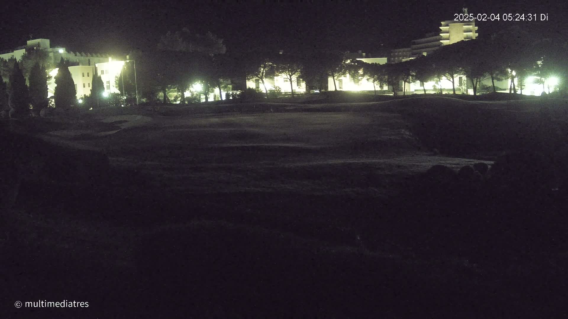 At night, dimly lit buildings are visible behind a dark, grassy area.