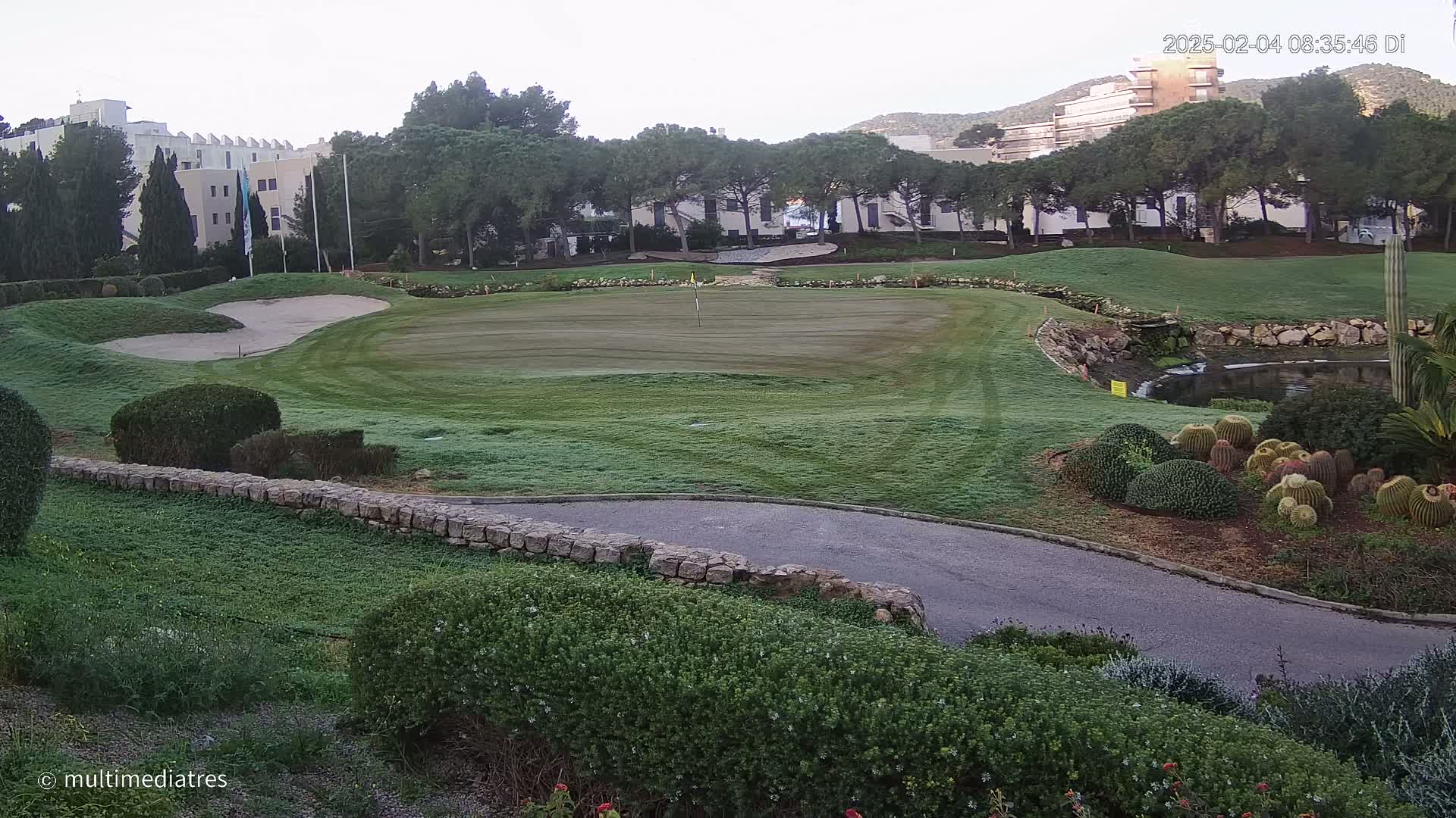 A golf course green with a sand trap and a small pond is visible, bordered by low stone walls and lush landscaping, under a mostly clear sky.