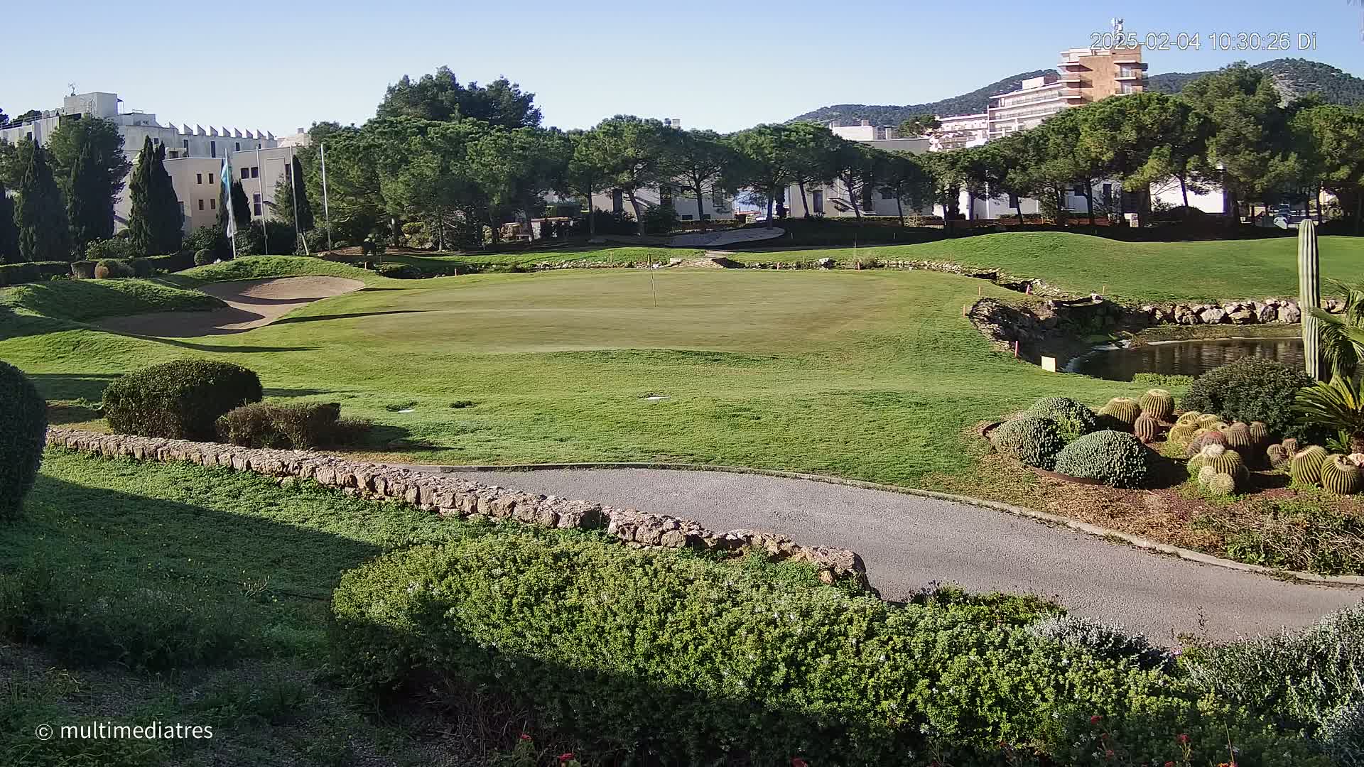 A golf course green with sand traps and a small pond is situated beside low buildings and a line of trees under a clear blue sky.