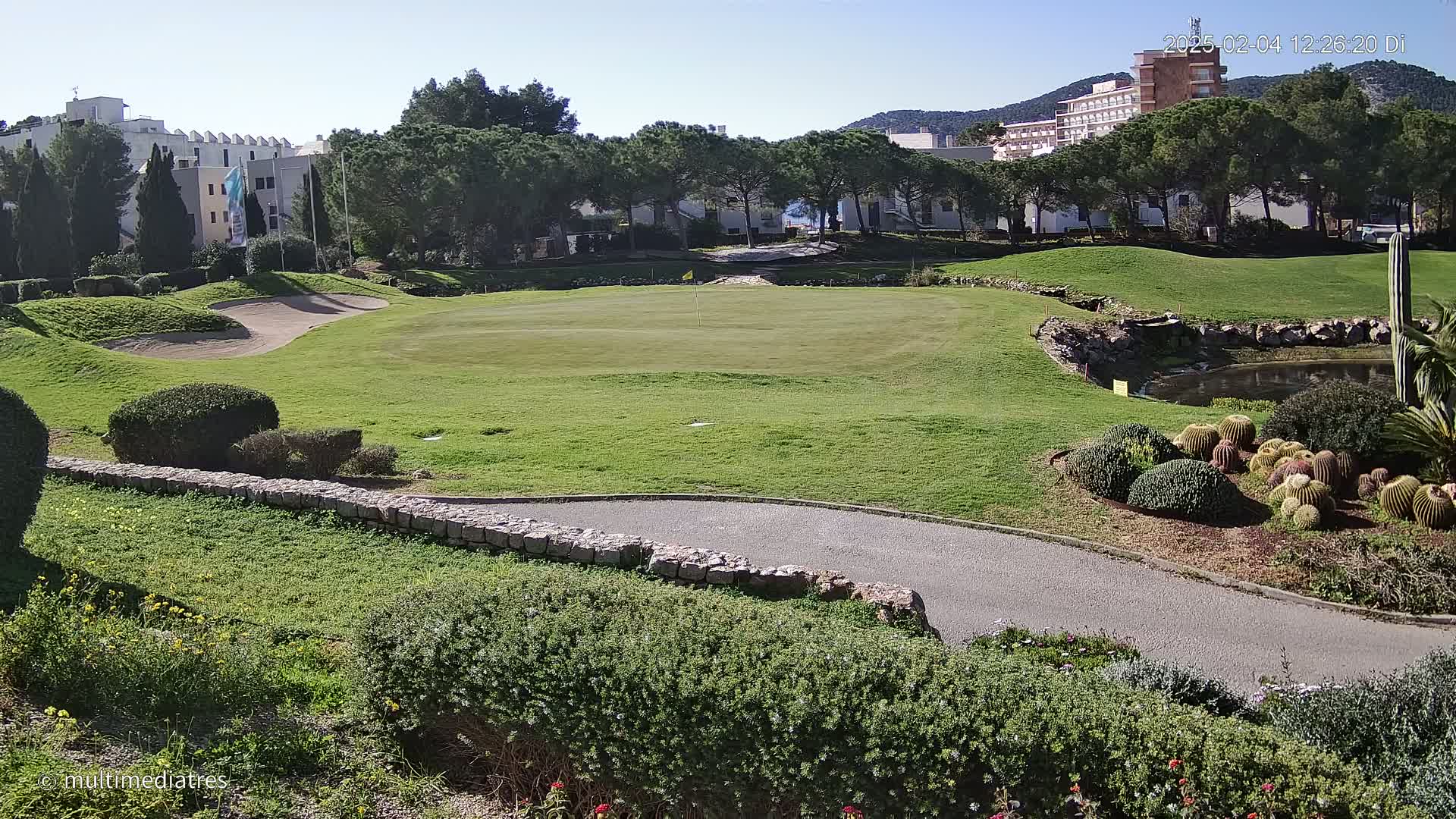 A sunny day overlooking a golf course green, sand trap, and small pond, with buildings and trees in the background.