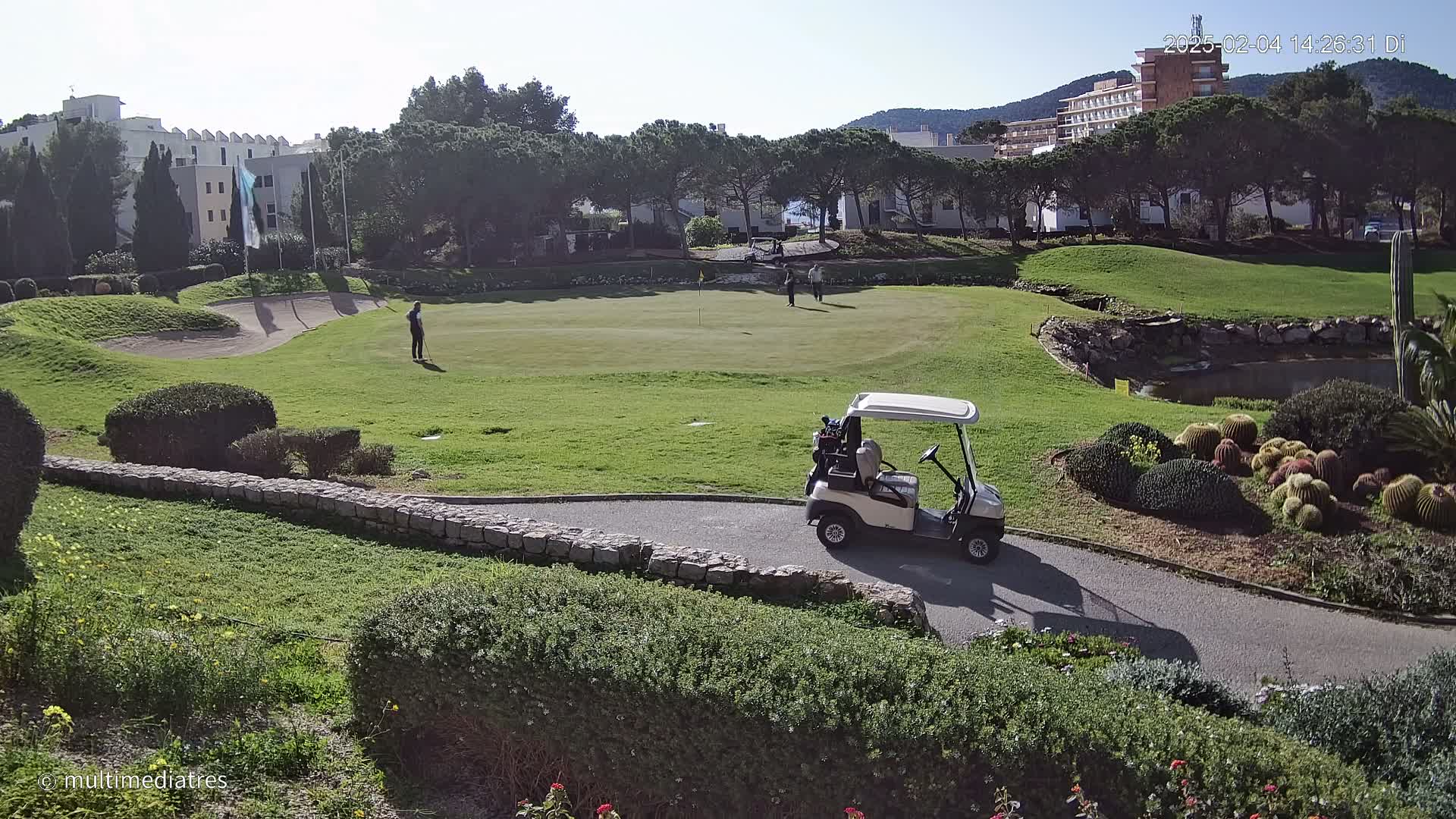 A golf course with several people playing golf, a golf cart on a path, and a building in the background on a sunny day.