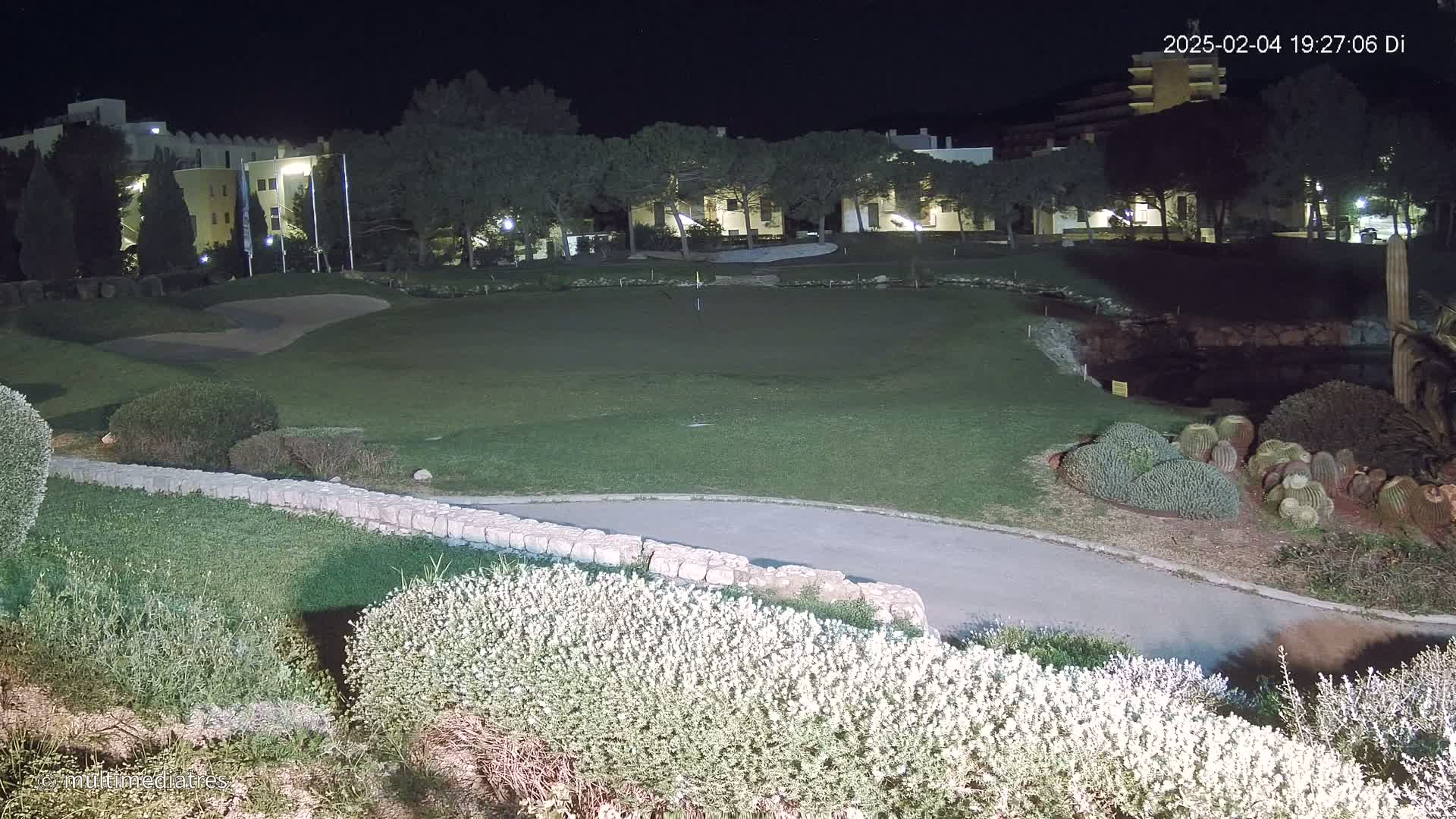 A nighttime view of a putting green illuminated by artificial light, adjacent to a building and landscaping with cacti.