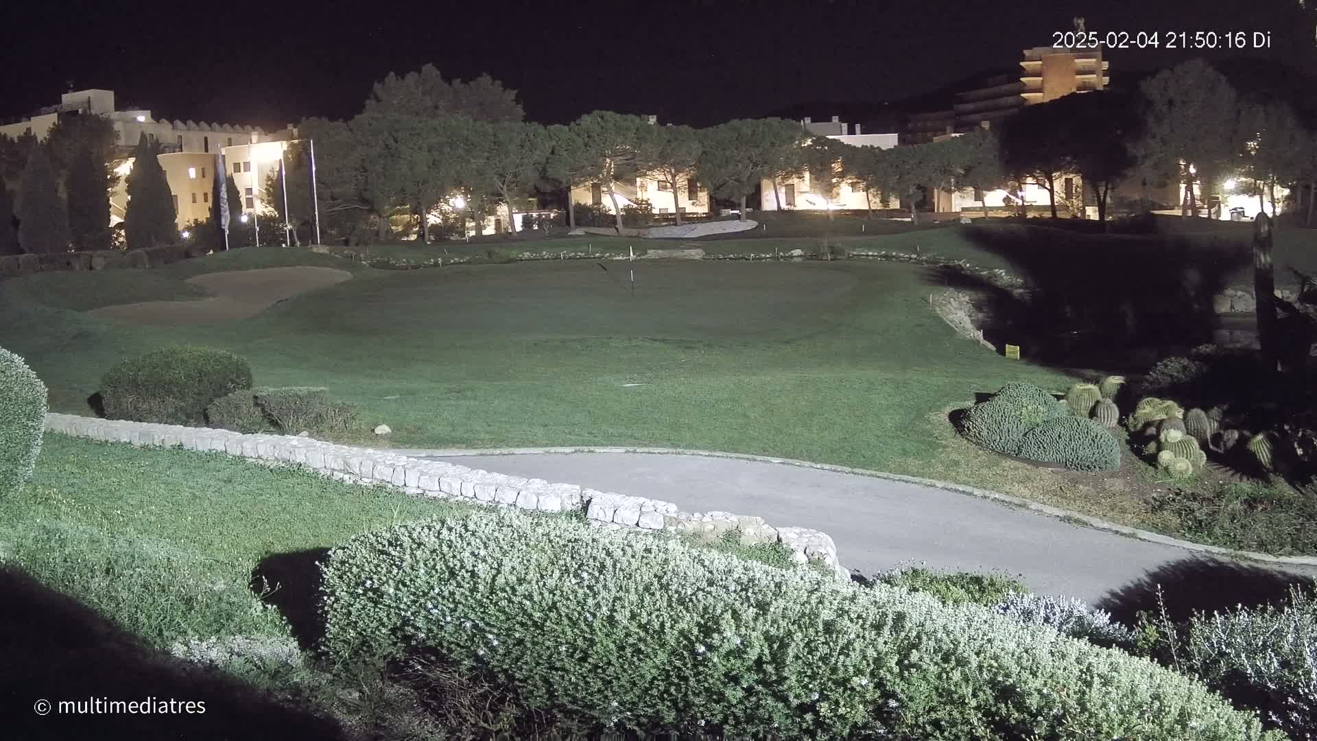 A nighttime view of a golf course green, illuminated by artificial light, with buildings and trees in the background.