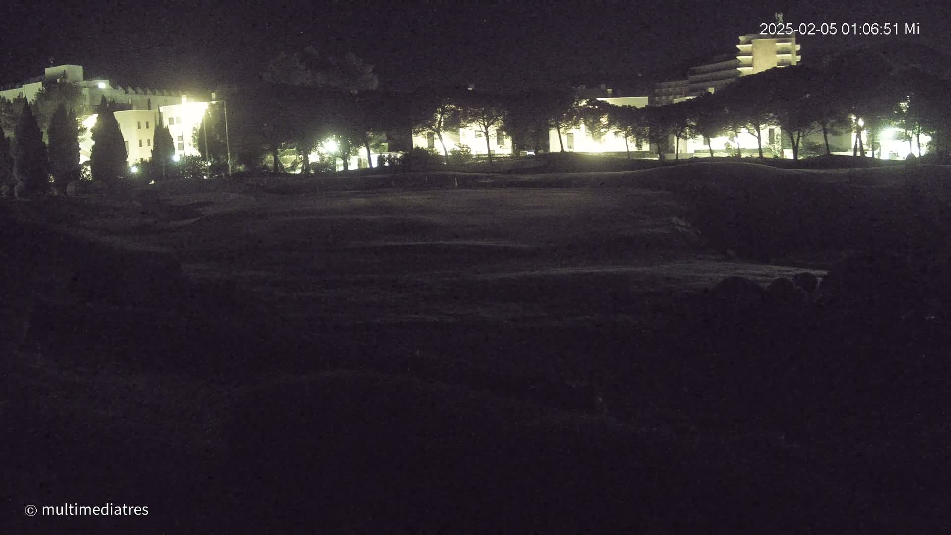 At night, illuminated buildings are visible beyond a dark, grassy expanse.