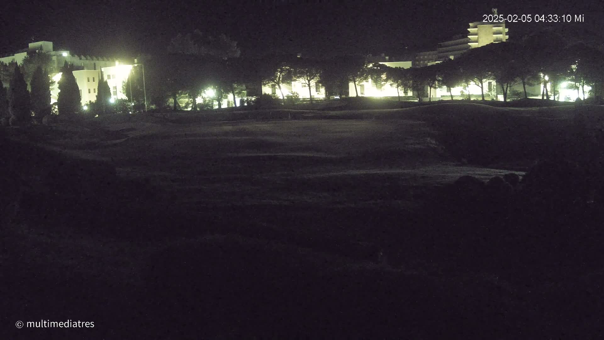 At night, illuminated buildings are partially visible behind a line of dark trees and a shadowed, grassy expanse.