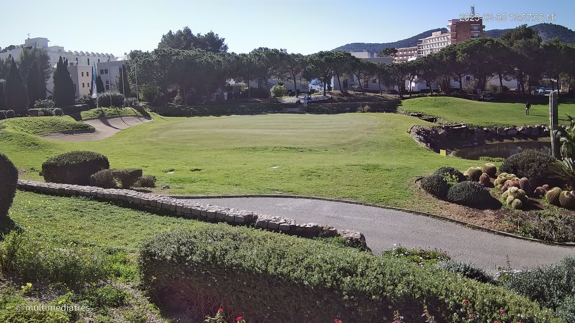 A sunny golf course with a green, sand trap, and a paved path is situated near buildings and lush green trees.