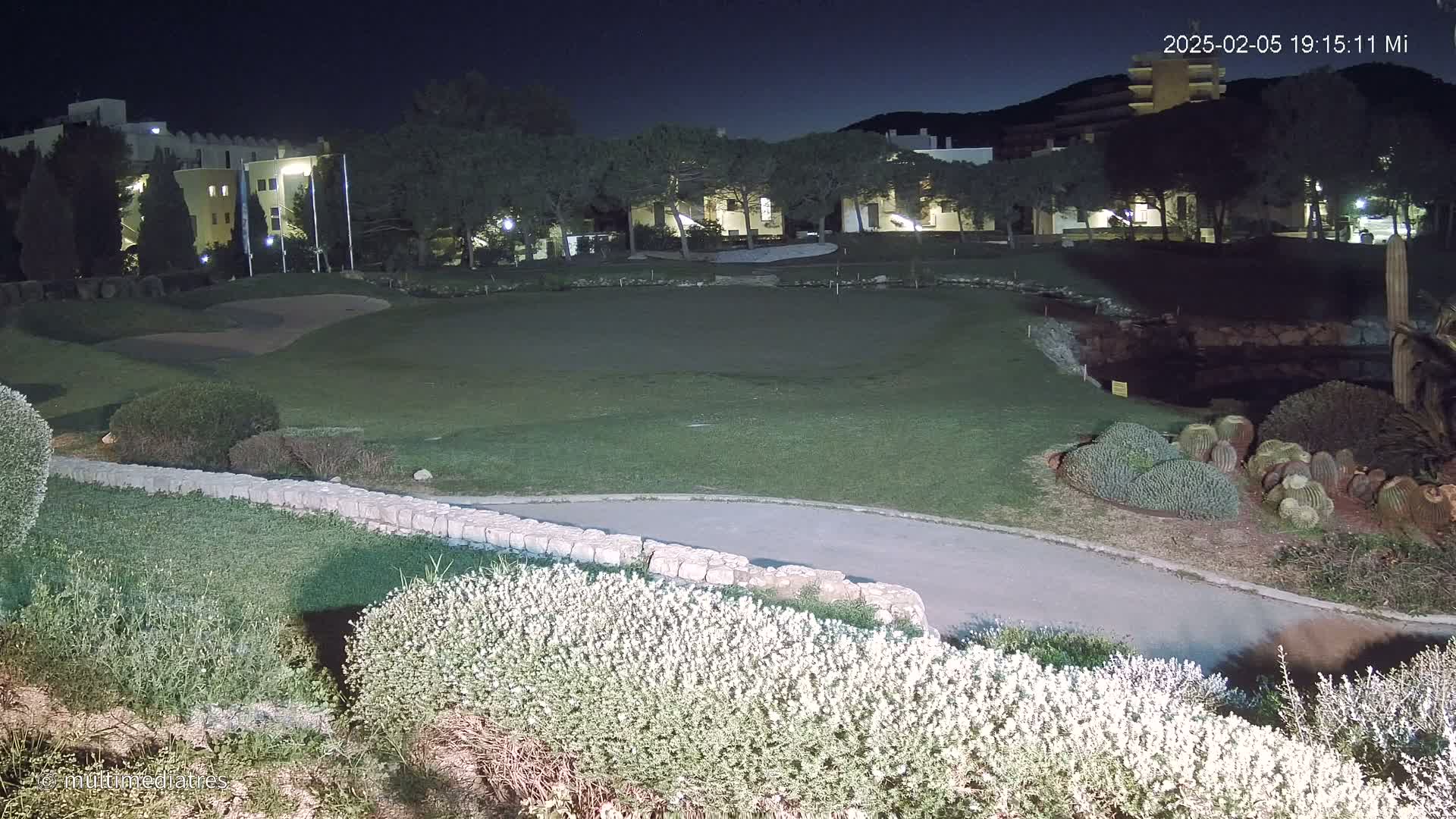 A nighttime view of a putting green and surrounding landscaping, with a building in the background under a dark sky.