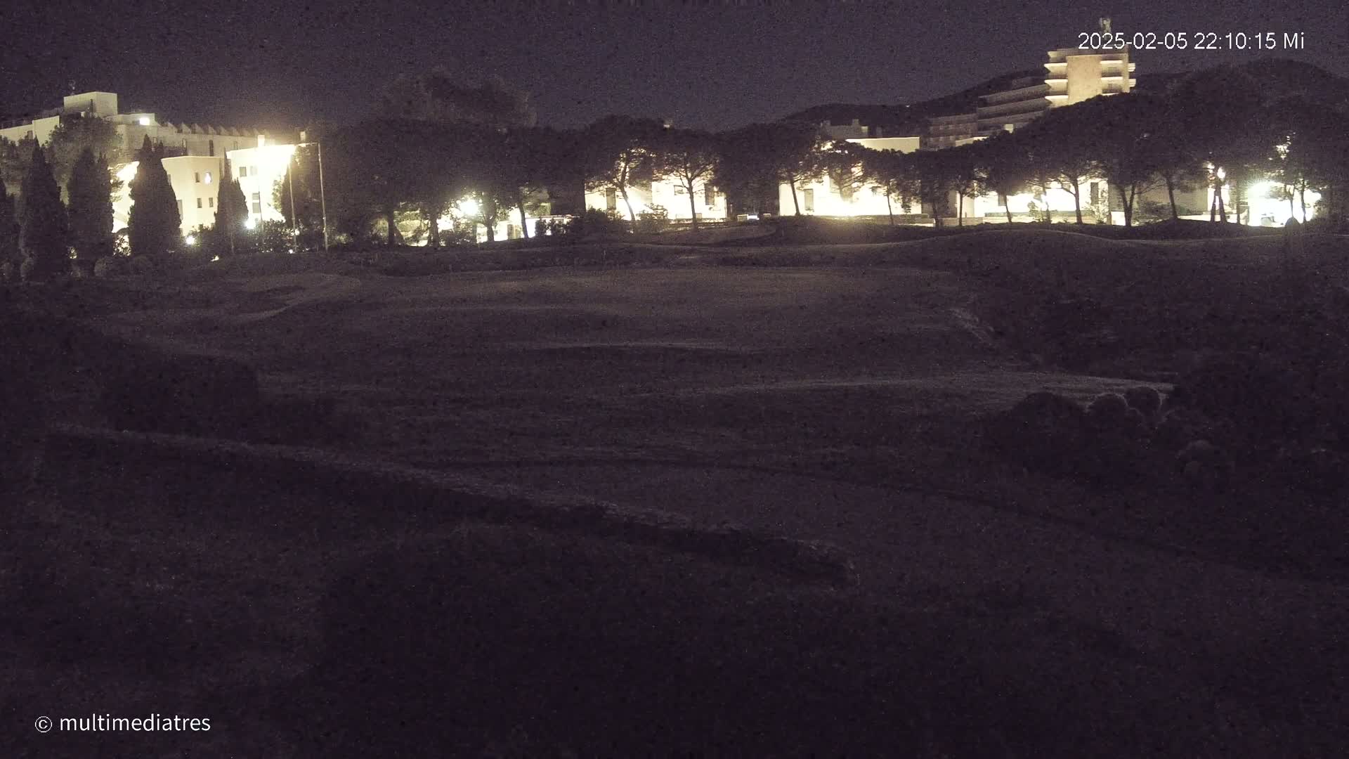 A nighttime, low-light view of a grassy area with illuminated buildings and trees in the background.