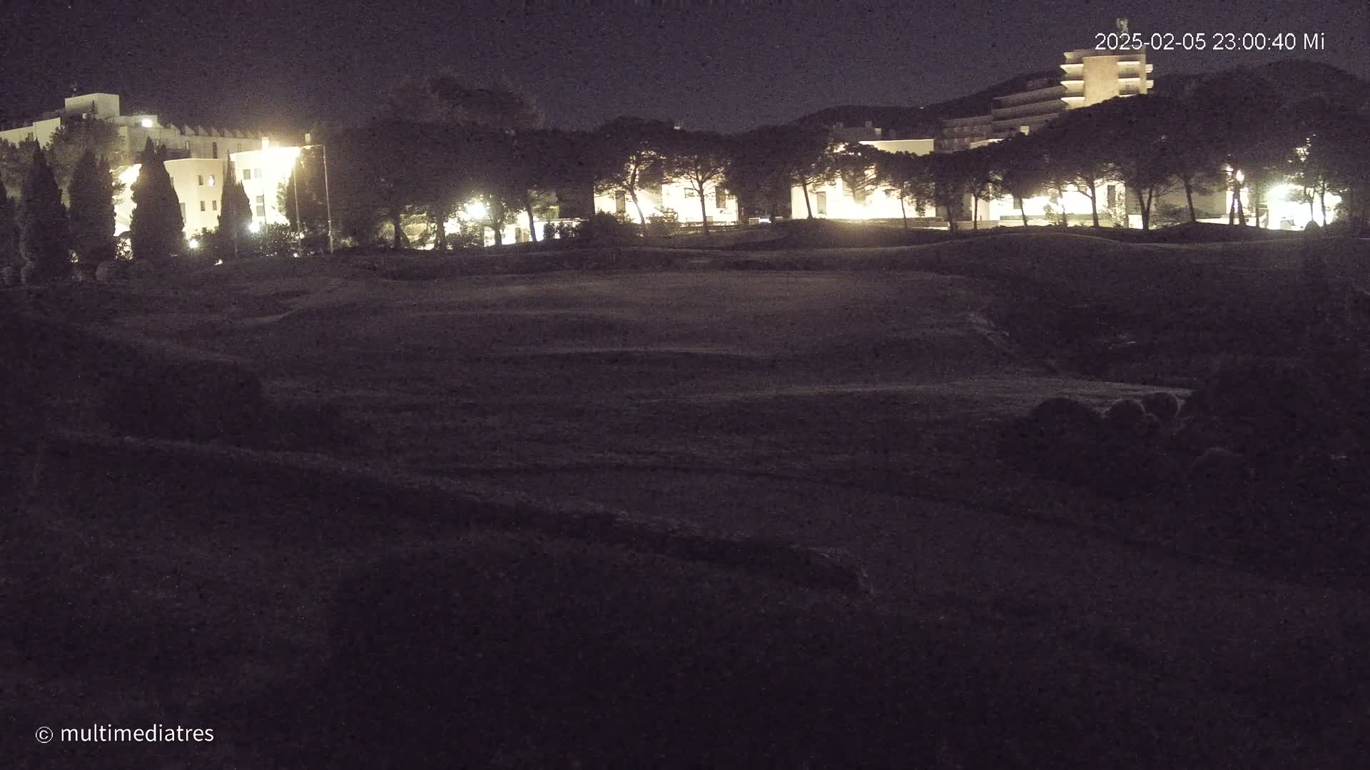 At night, a dark, grassy area stretches before several illuminated buildings surrounded by trees.
