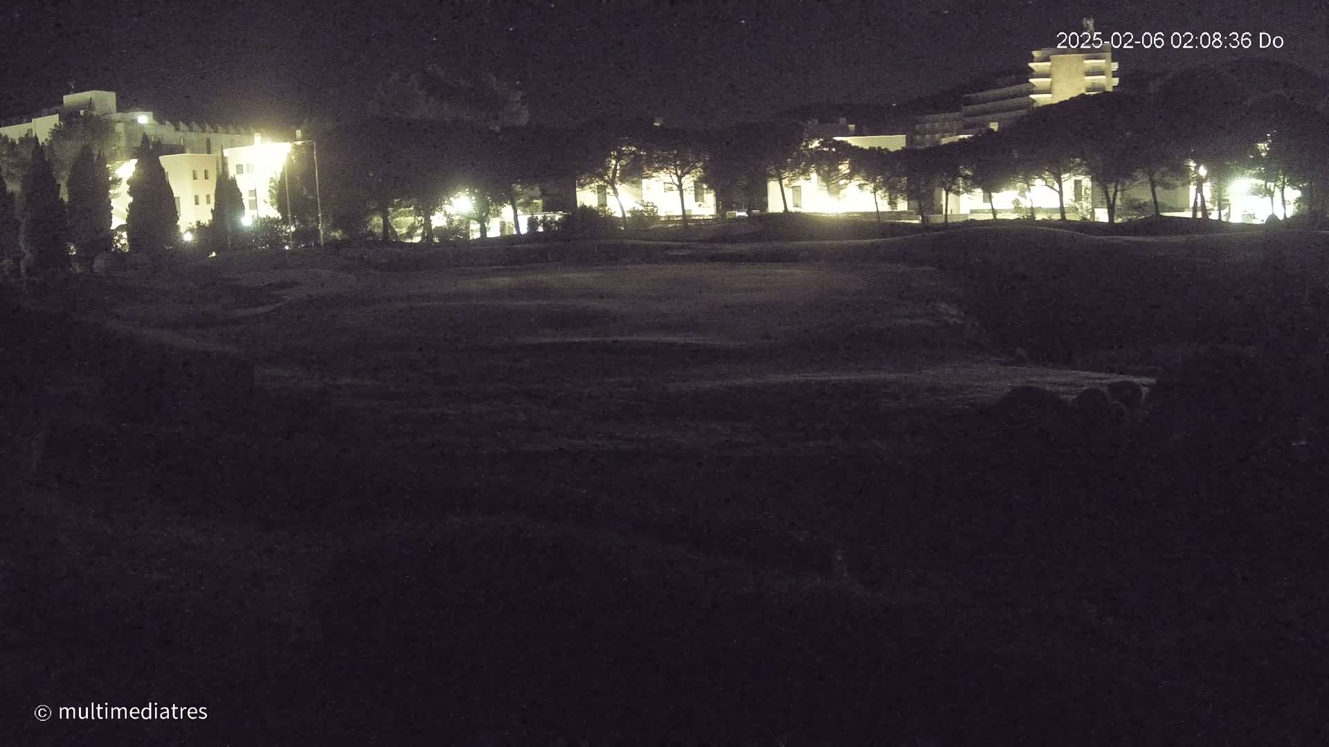 A nighttime view of a dark, grassy area with illuminated buildings and trees in the background under a clear sky.