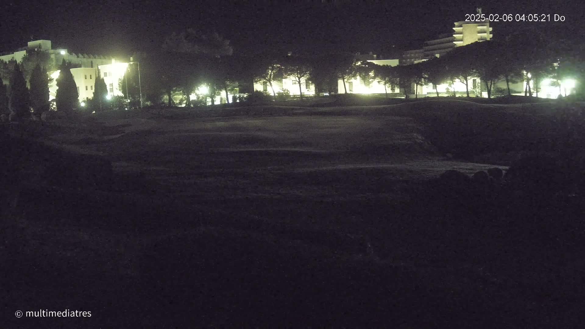 A nighttime view of a dark, grassy area with illuminated buildings and trees in the background.