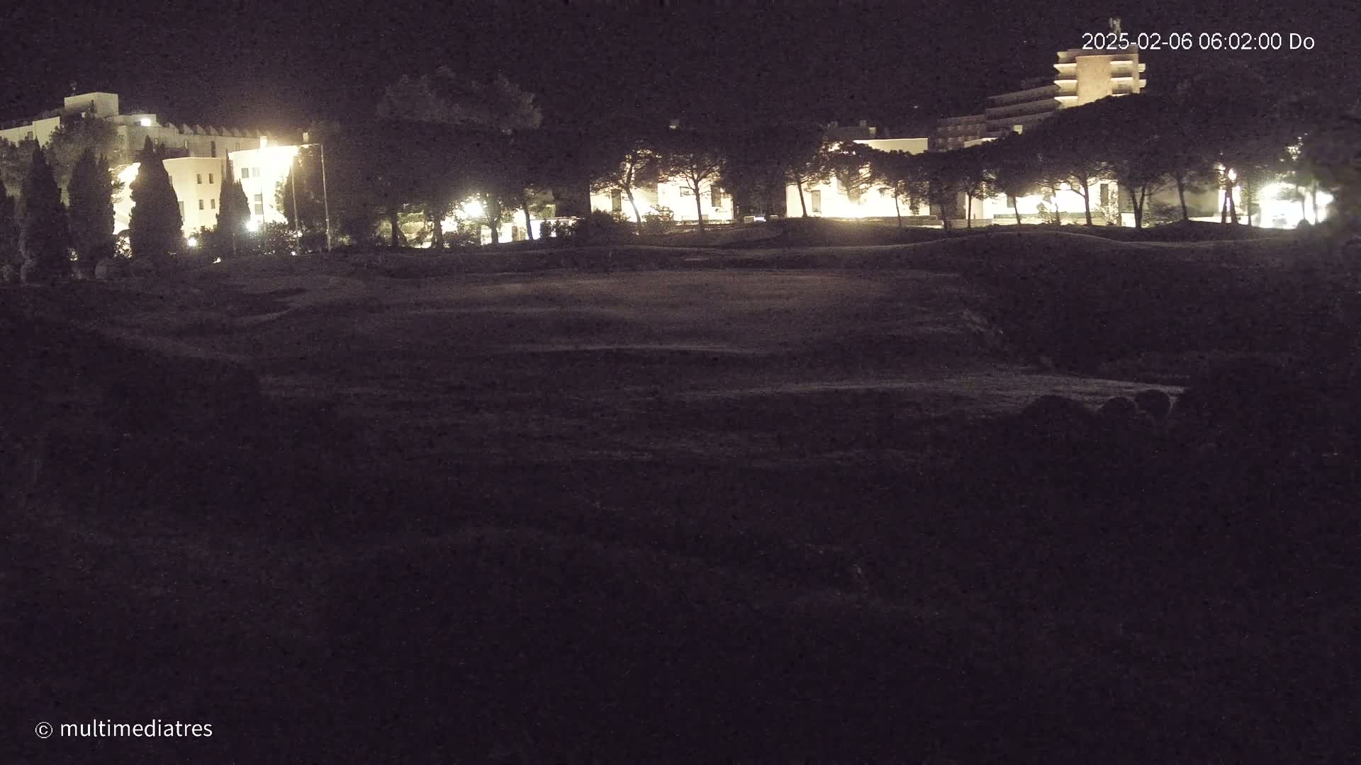 At night, illuminated buildings are partially obscured by a row of dark trees in front of a dark, grassy expanse.
