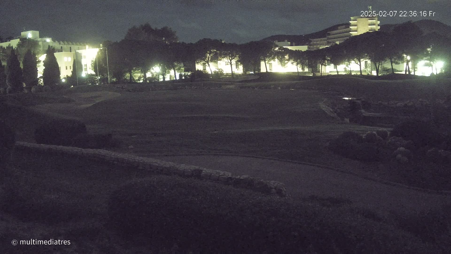 A nighttime view of a grassy area with buildings and trees illuminated in the background under a partly cloudy sky.