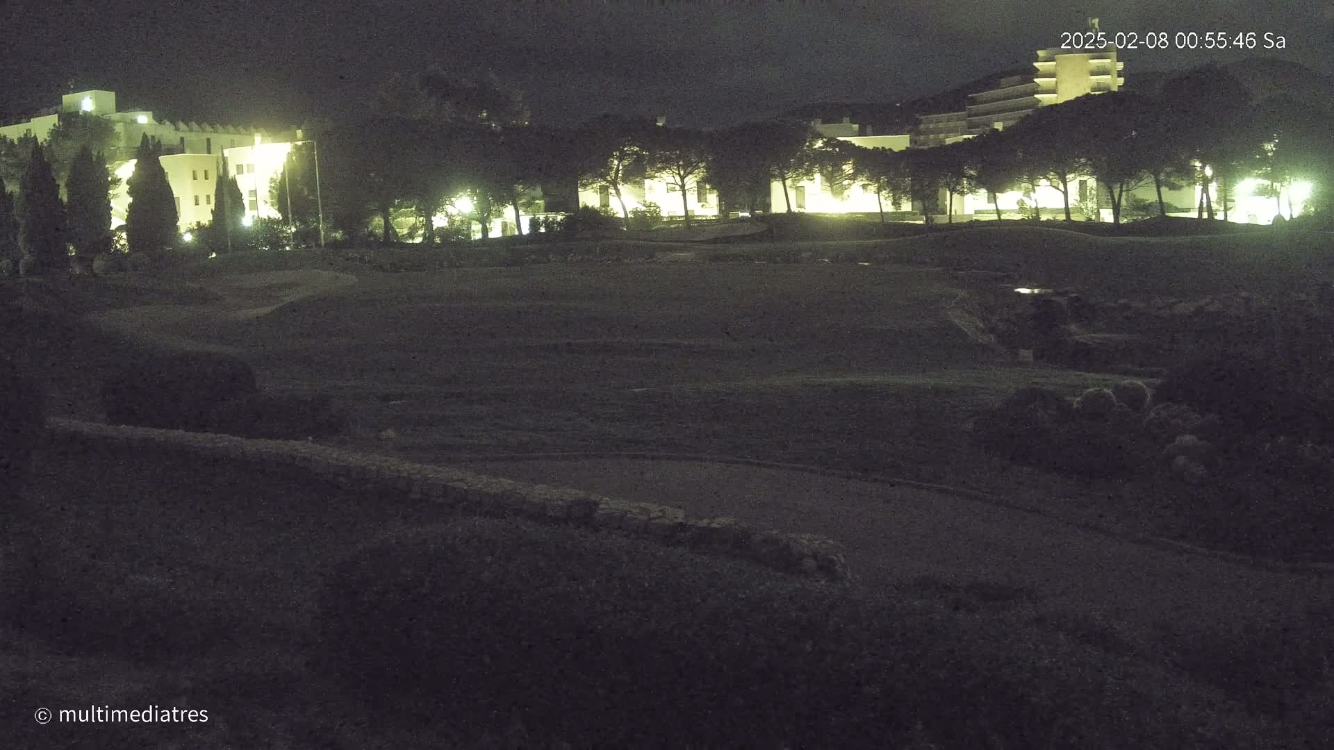 A nighttime view of a grassy field with dimly lit buildings in the background under a dark sky.