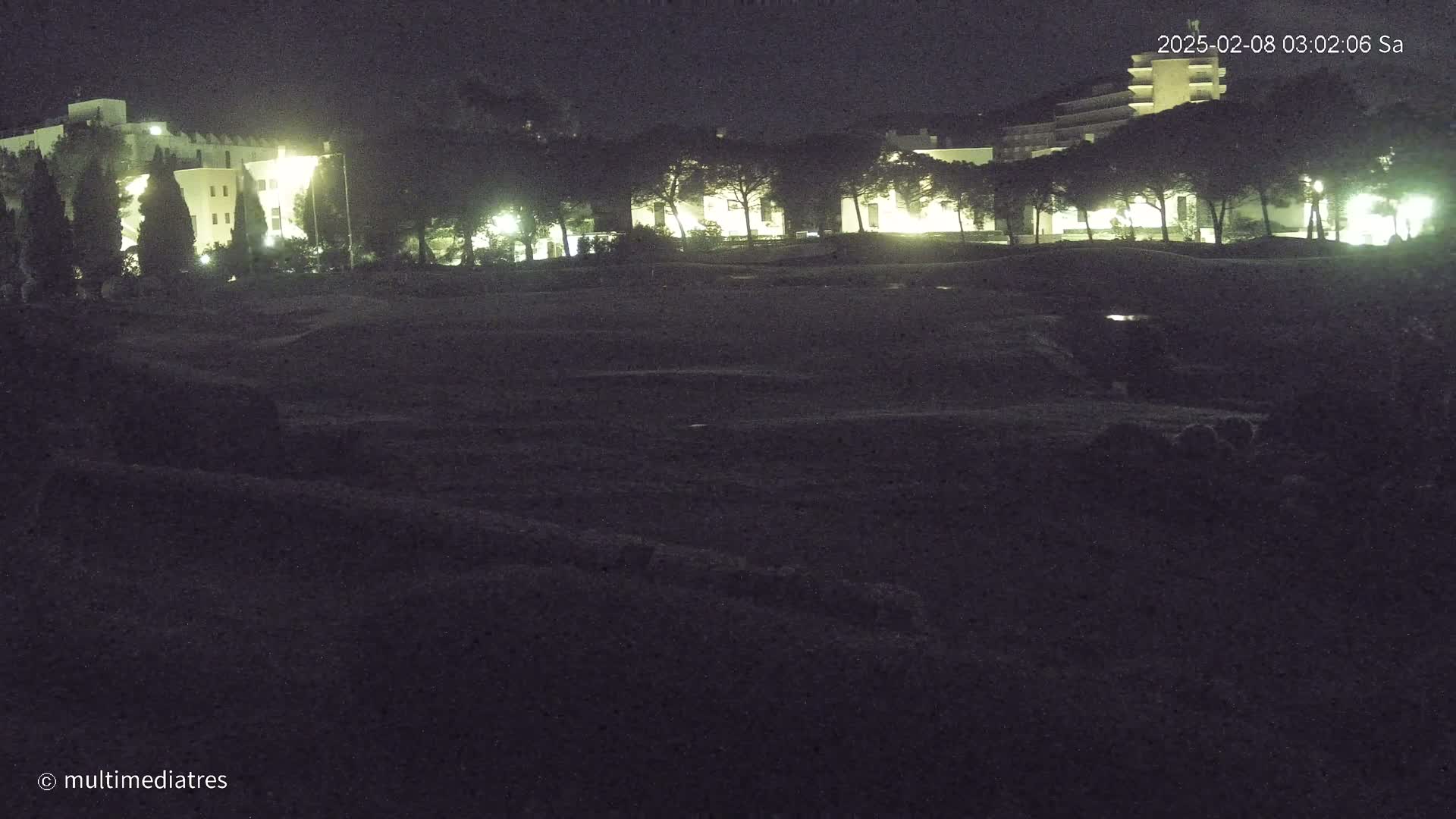 At night, illuminated buildings are visible behind a dark, flat expanse of land with a few trees.