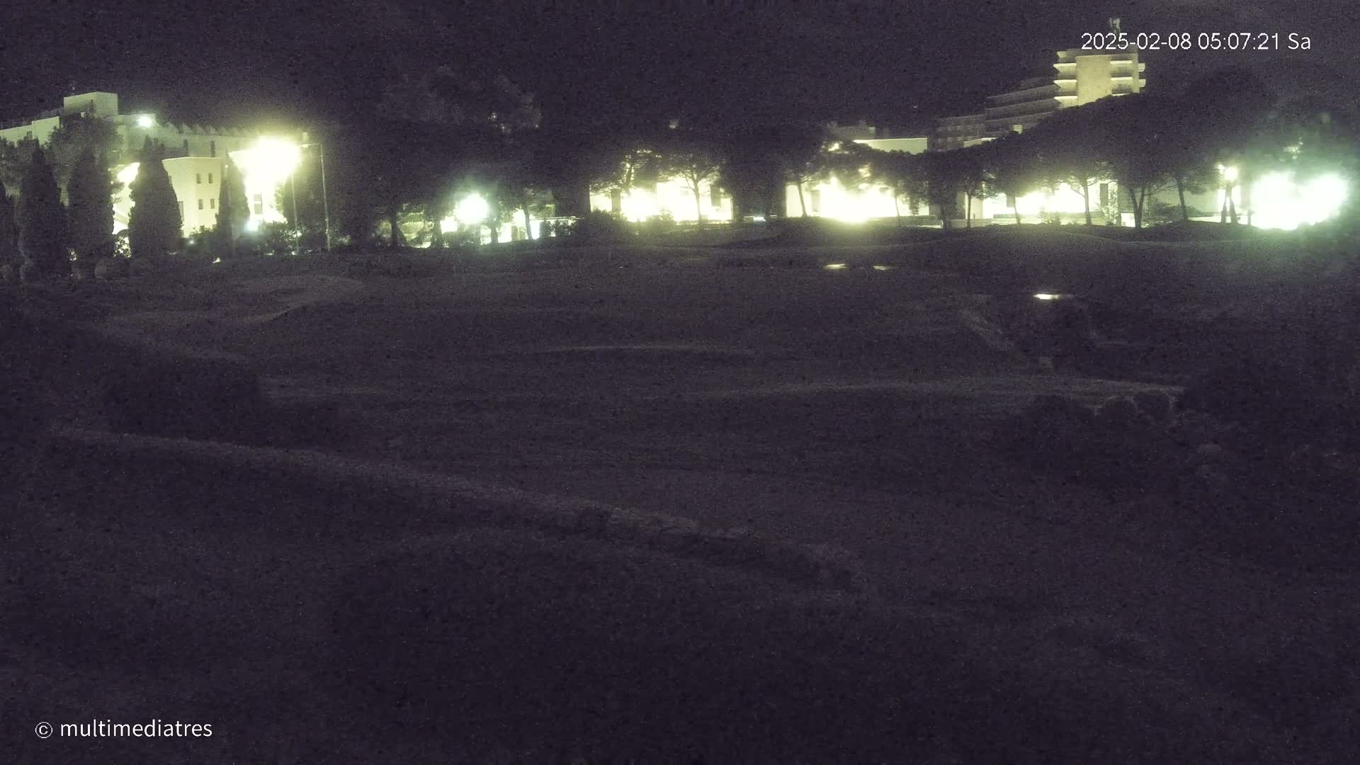 A dark, nighttime scene shows a flat, dark expanse of ground in front of dimly lit buildings and trees.