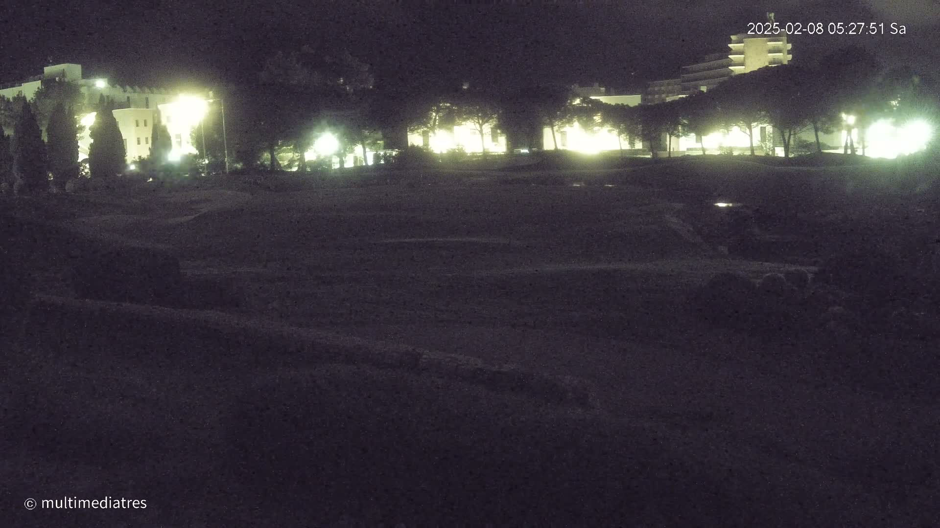 At night, illuminated buildings and trees are visible across a dark, seemingly empty field.