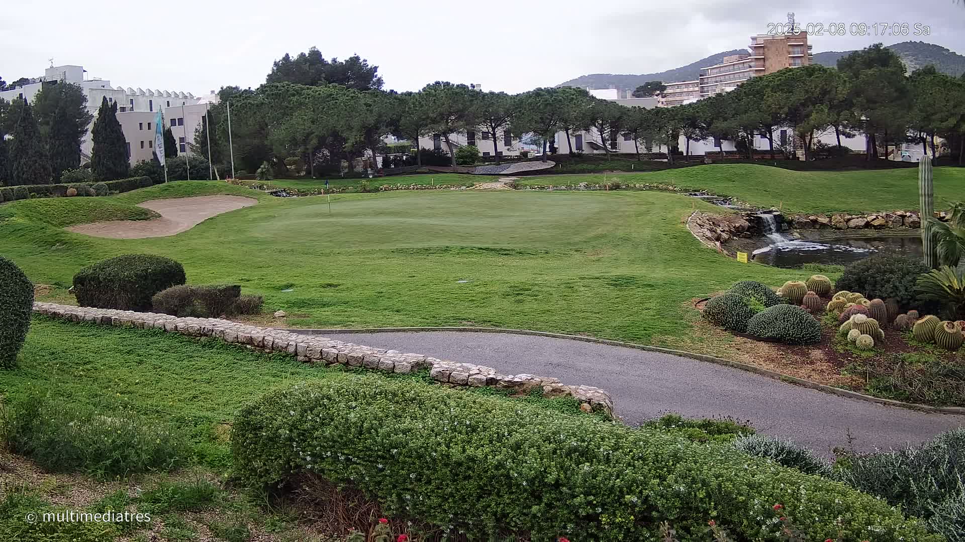 A golf course green with a sand trap, a small waterfall and pond, and lush landscaping under an overcast sky.