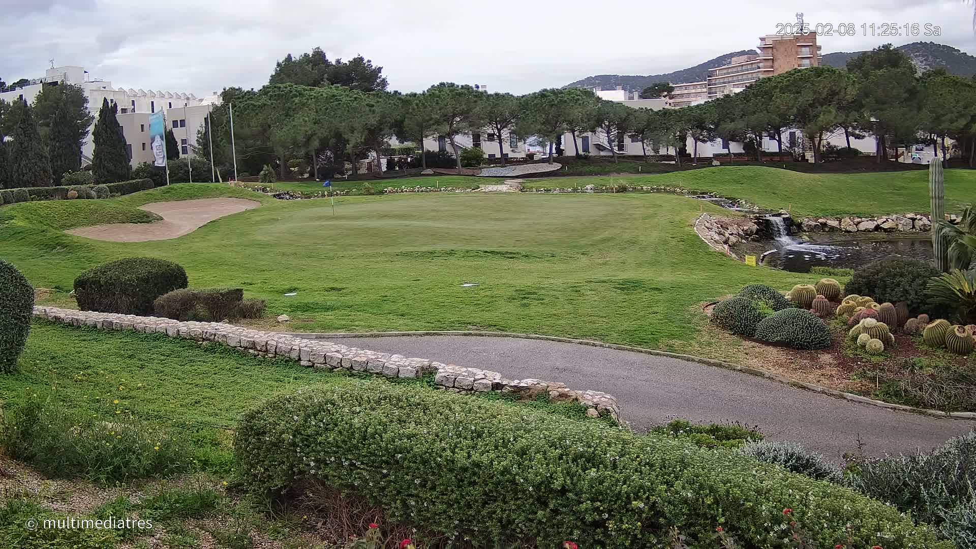 A golf course green with a sand trap, a small waterfall, and a collection of cacti, all under a cloudy sky.