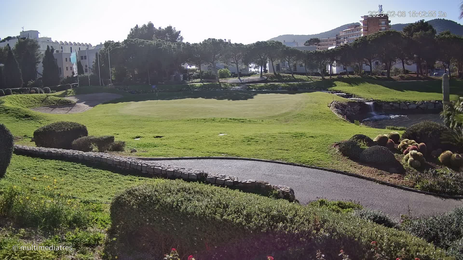 A sunny day reveals a golf course with a green, a small waterfall, and a low stone wall curving alongside a paved path, all set against a backdrop of buildings and trees.
