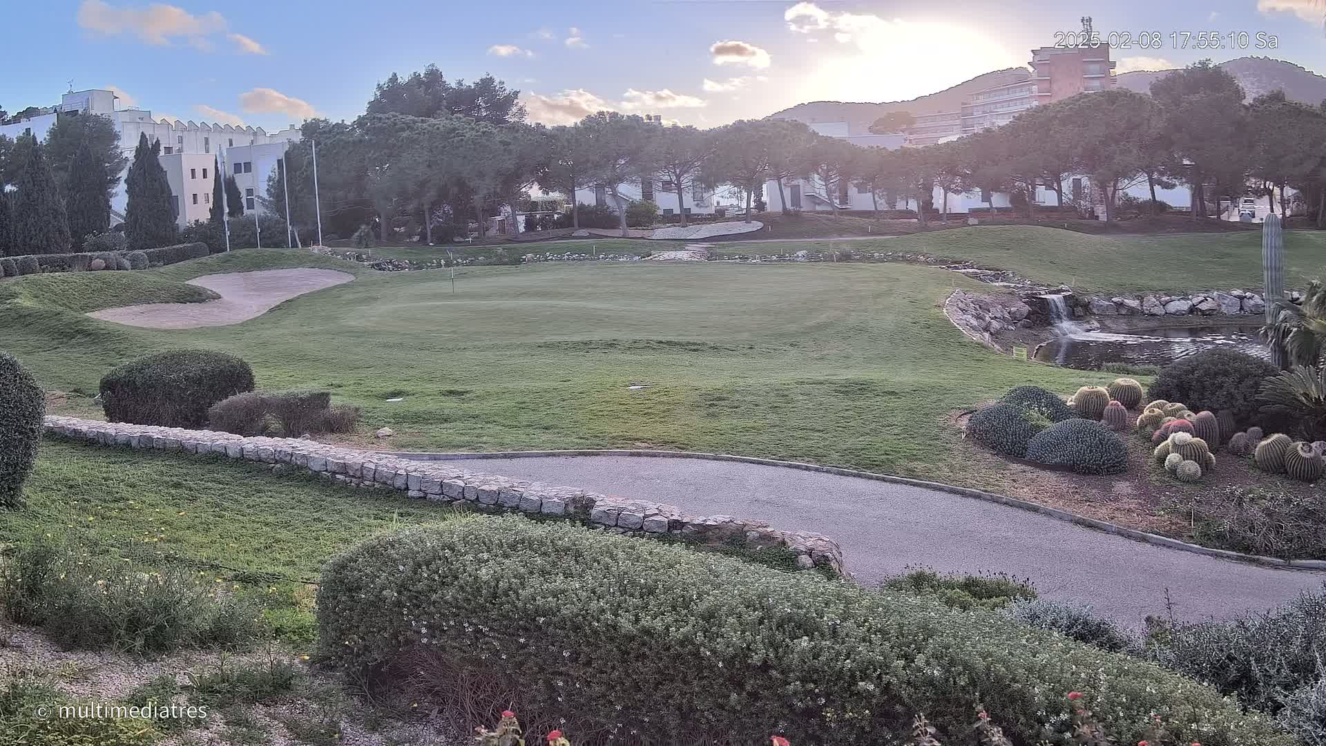 A golf course green with a sand trap, a small pond with a waterfall, and a collection of cacti under a partly cloudy sky.
