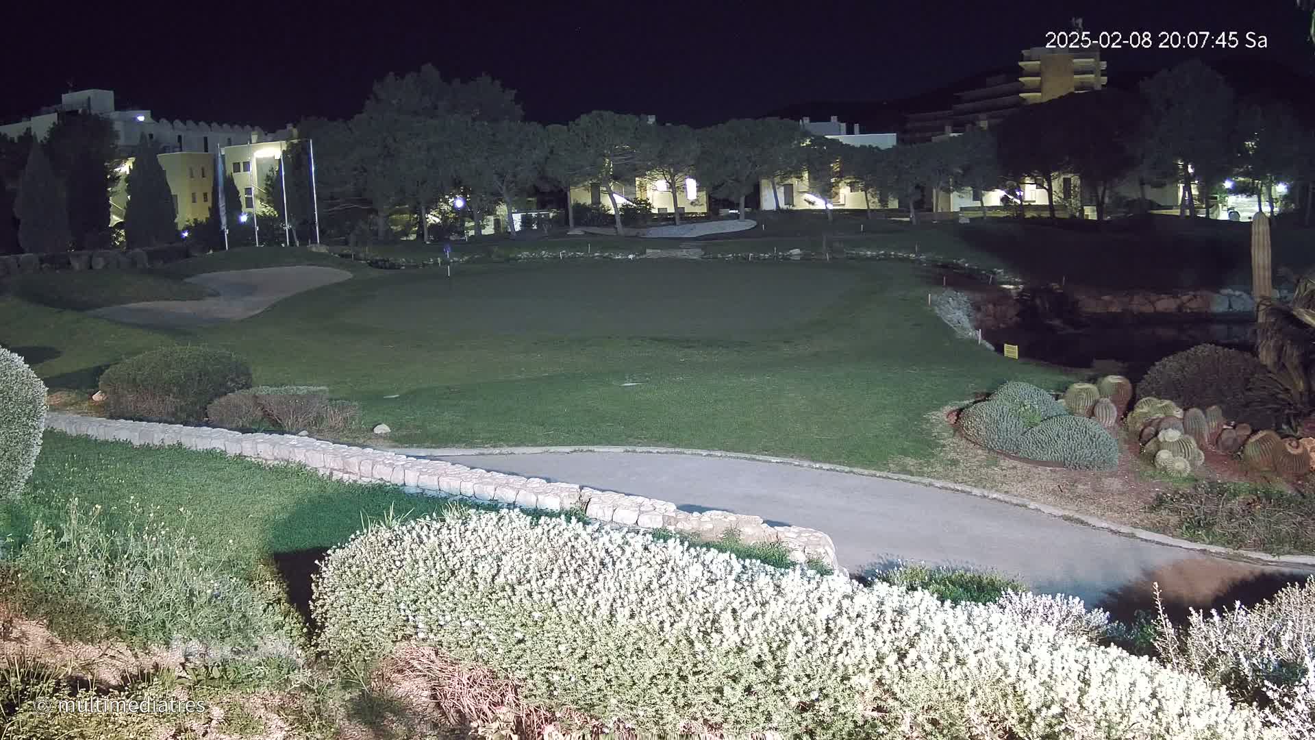 A nighttime view of a putting green and surrounding landscaping, including low shrubs, a stone wall, and a building in the background, illuminated by artificial light.
